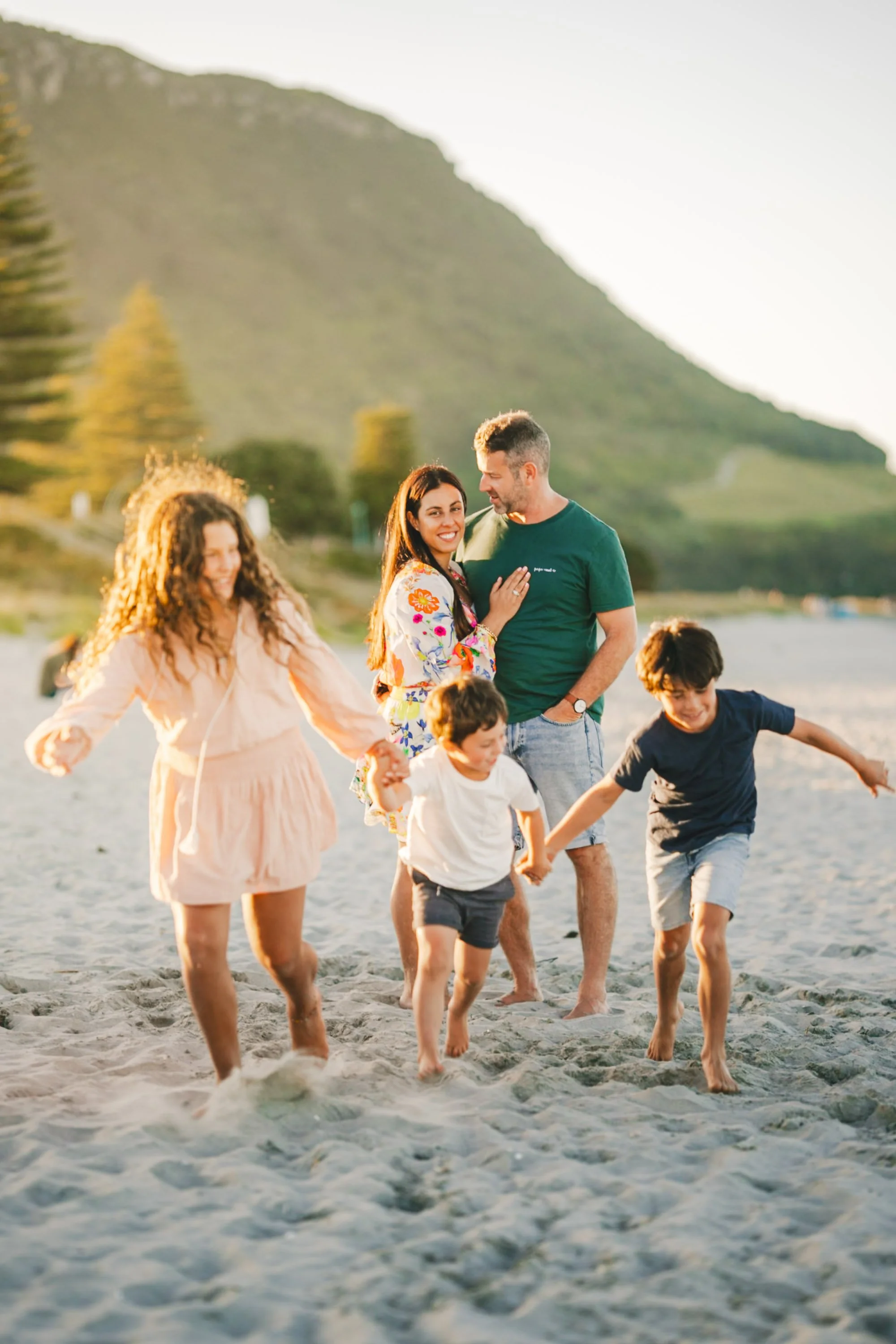 A family of five enjoying a day at the beach, walking and playing in the sand near the water with a mountain and trees in the background in warm sunset lighting.