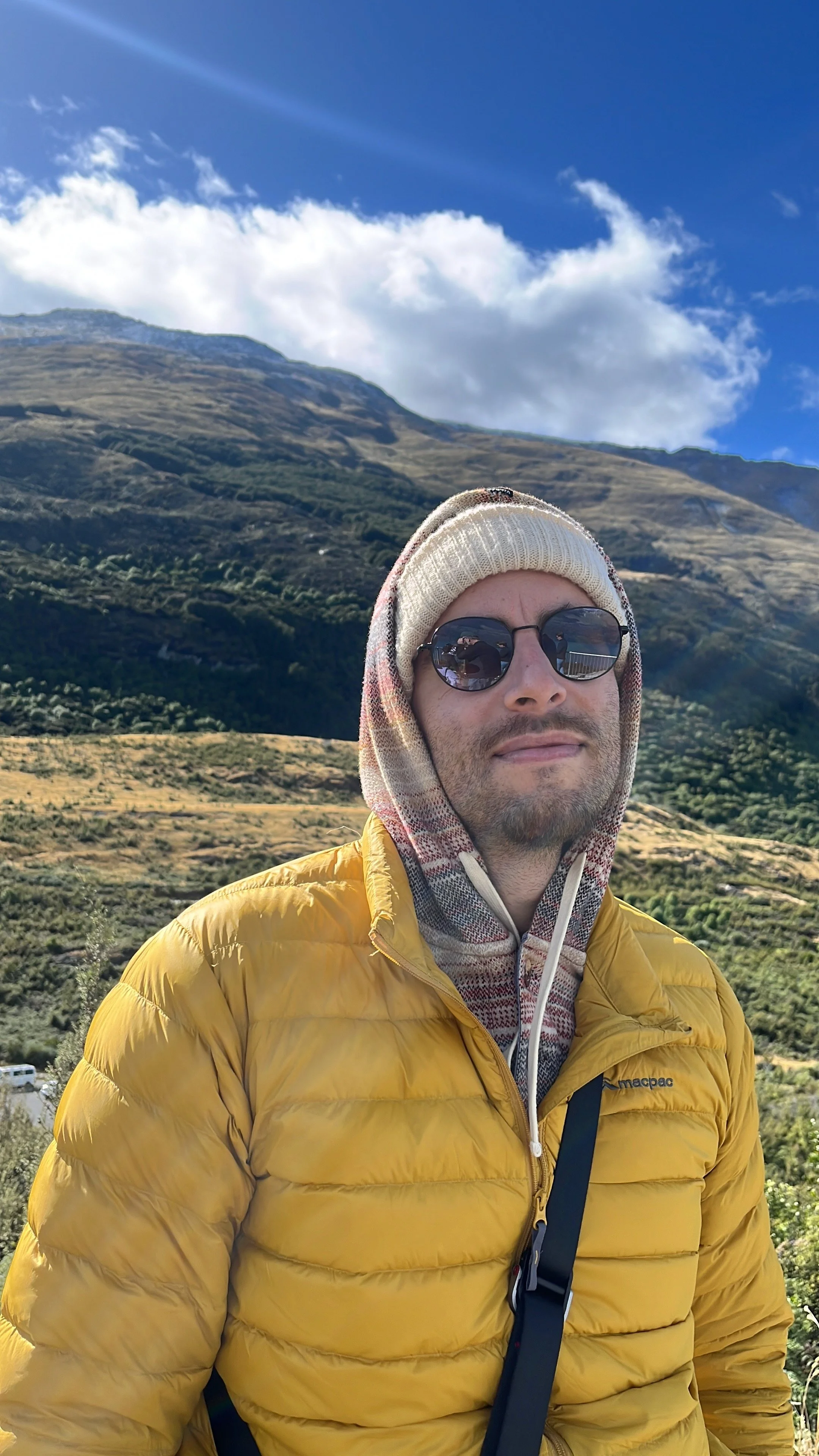 Man wearing sunglasses, a beige beanie, and a yellow puffy jacket, standing outdoors in a mountainous landscape with green hills, a blue sky, and white clouds.