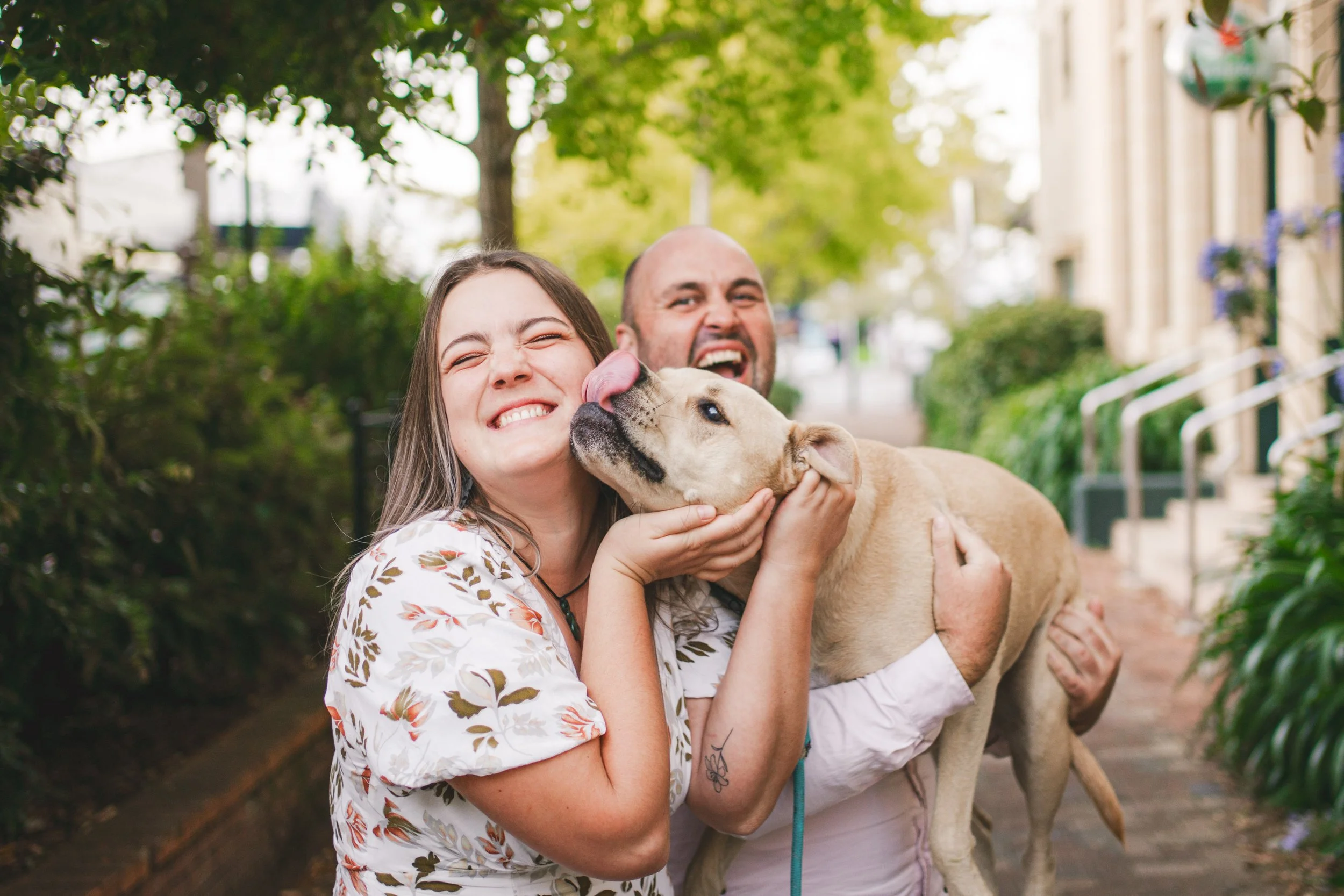 A smiling woman and a man with a dog, outdoors on a city street.