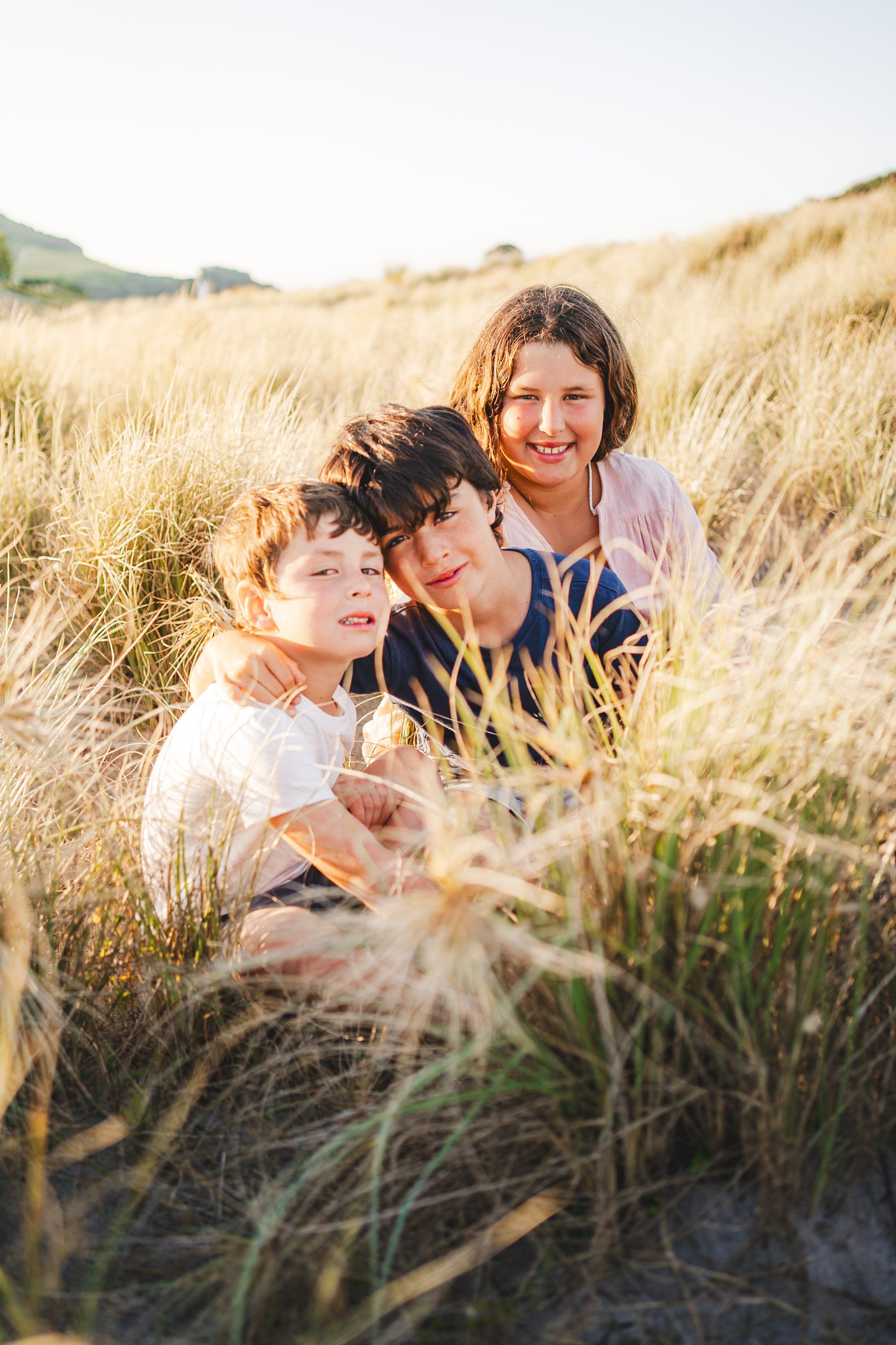 Three children sitting in tall grass on a sunny day, smiling at the camera.