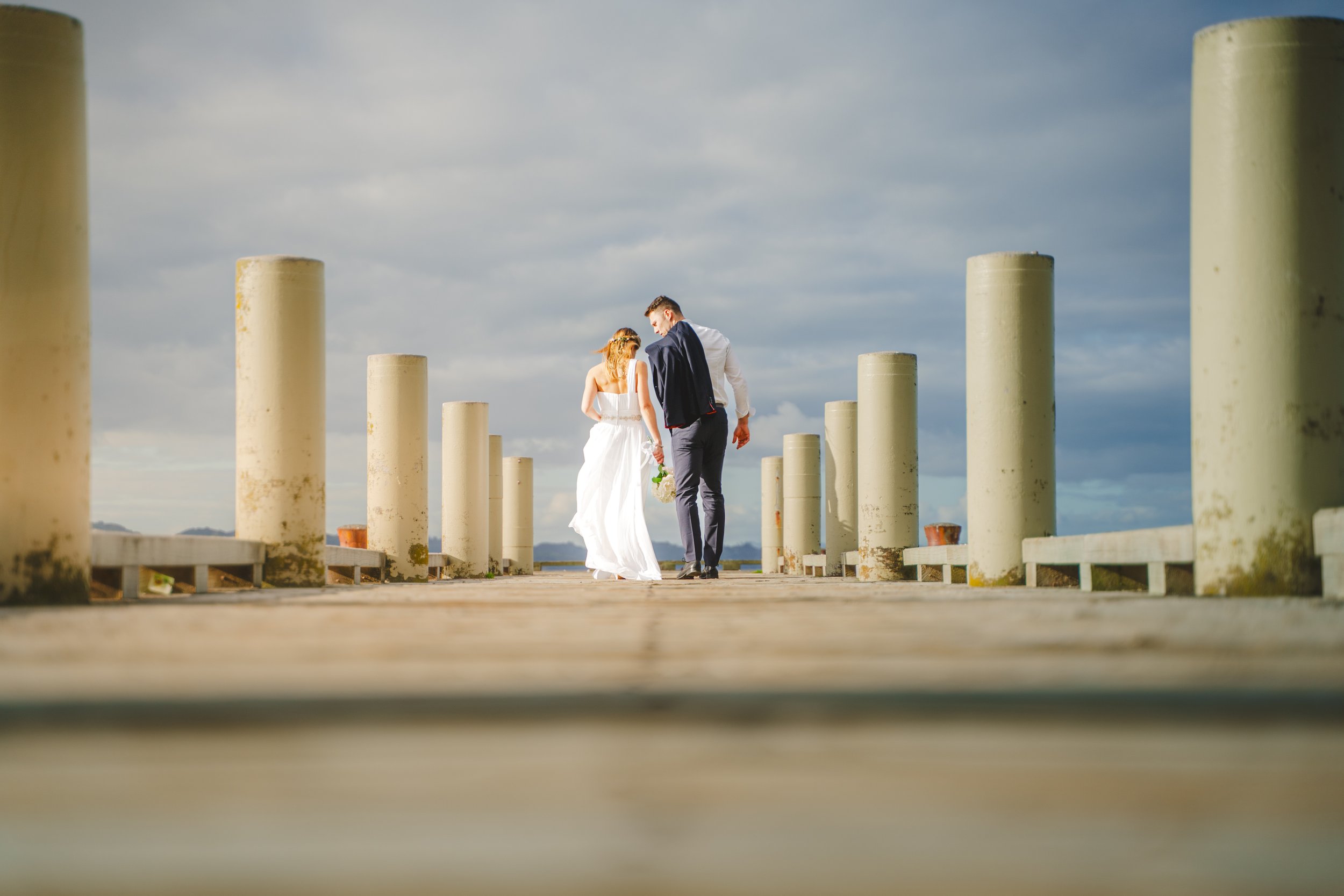 A bride and groom walking on a wooden pier with white pilings, holding hands, under a cloudy sky.