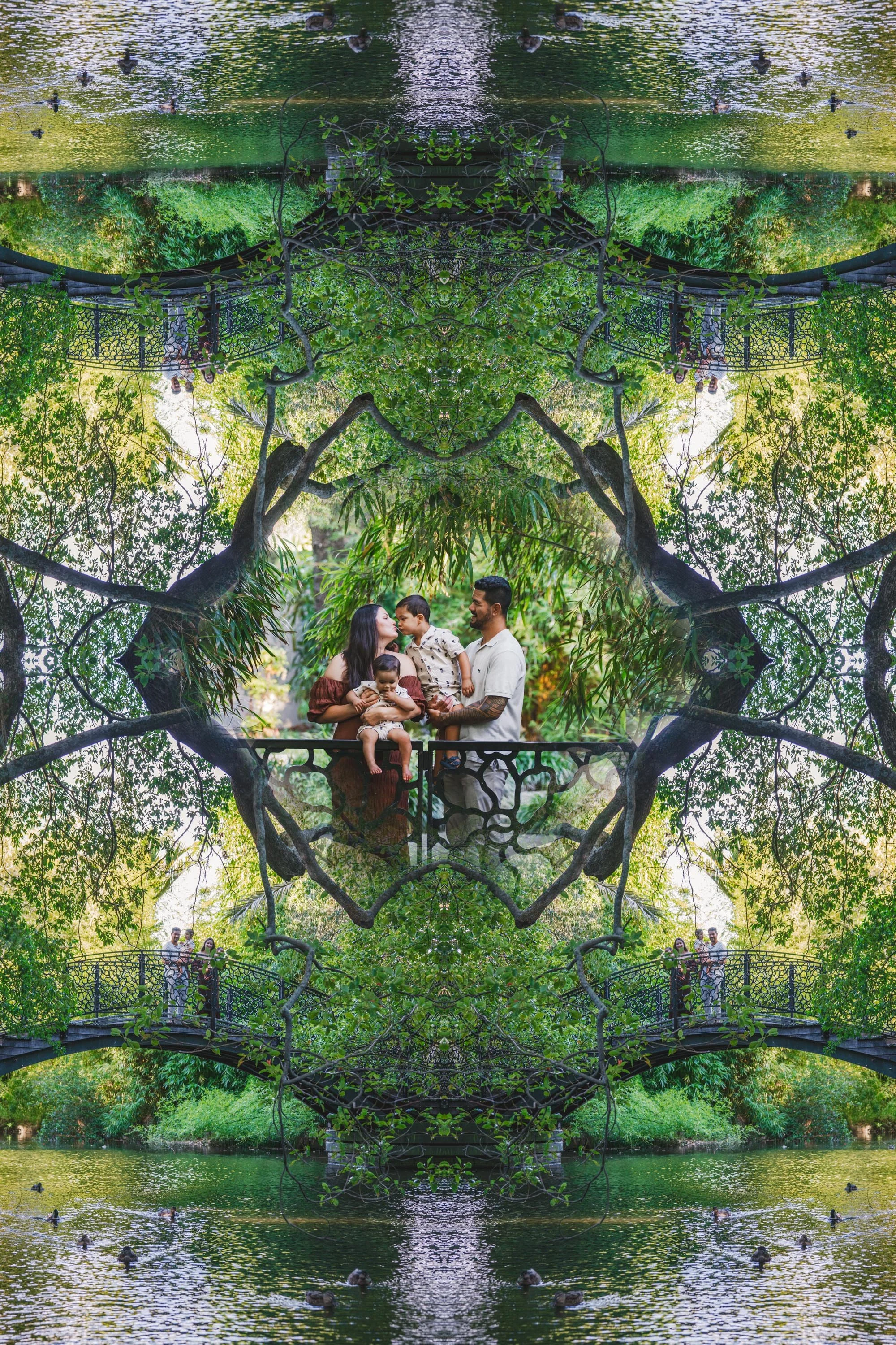 A family of four stands on a bridge in a lush green park, reflected symmetrically in a mirrored image, with trees, water, and ducks.