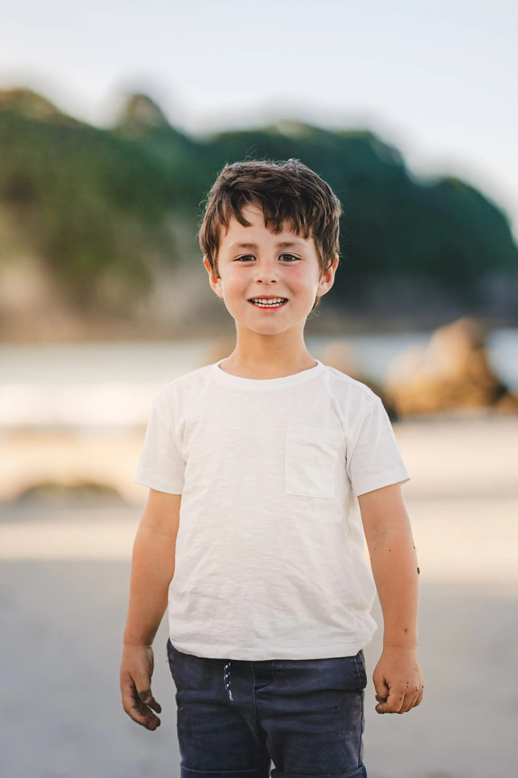 A young boy with brown hair and a white t-shirt, smiling at the camera on a sandy beach with rocks and trees in the background.
