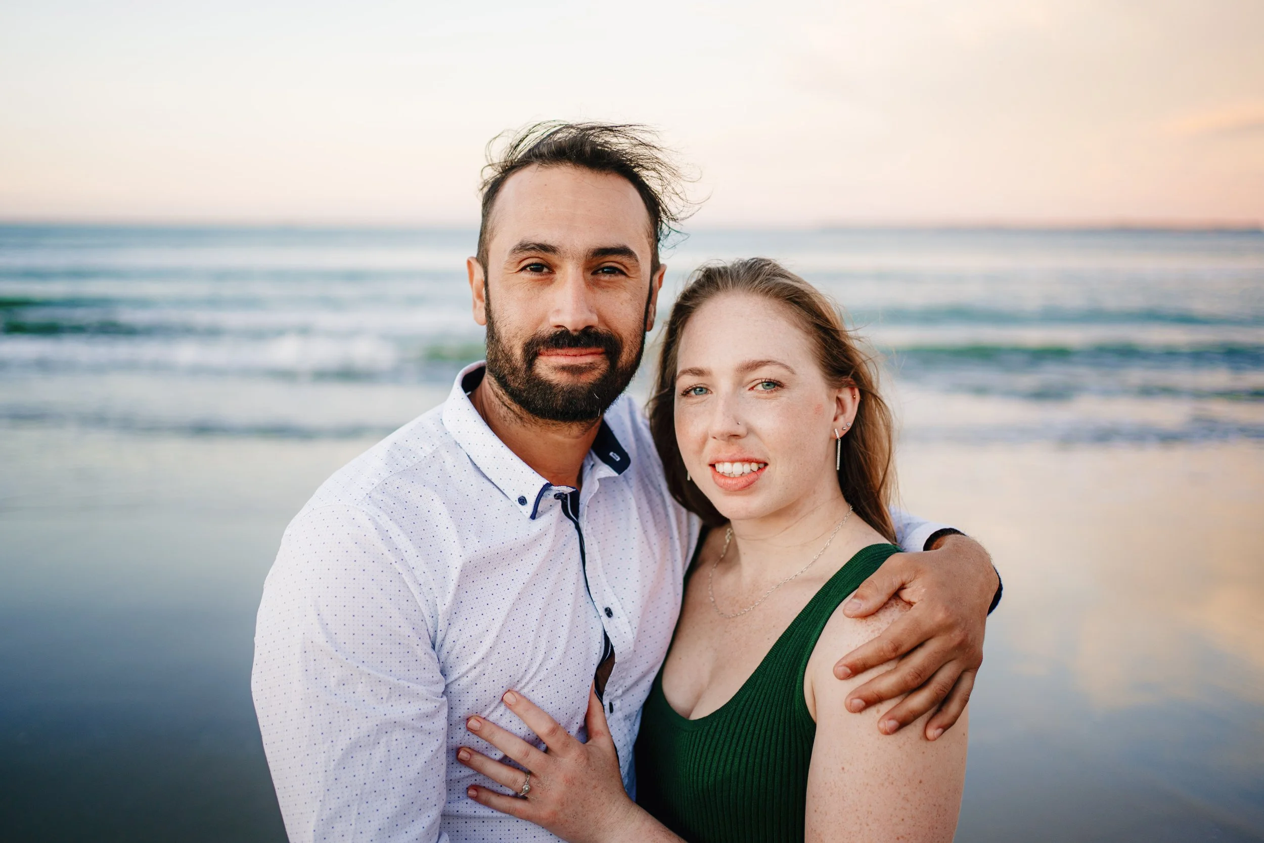 A couple standing close together on the beach at sunset, with ocean waves in the background.
