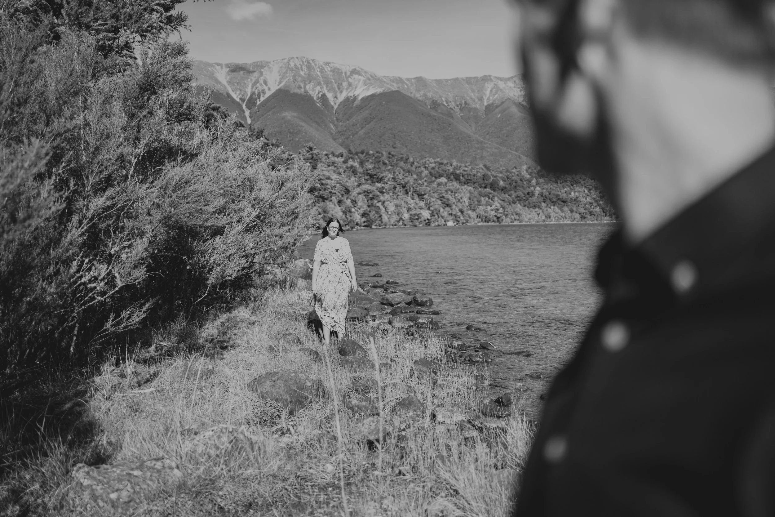 A woman walking along a lakeshore with mountains in the background, and a man partially visible in the foreground on the right side of the photo.