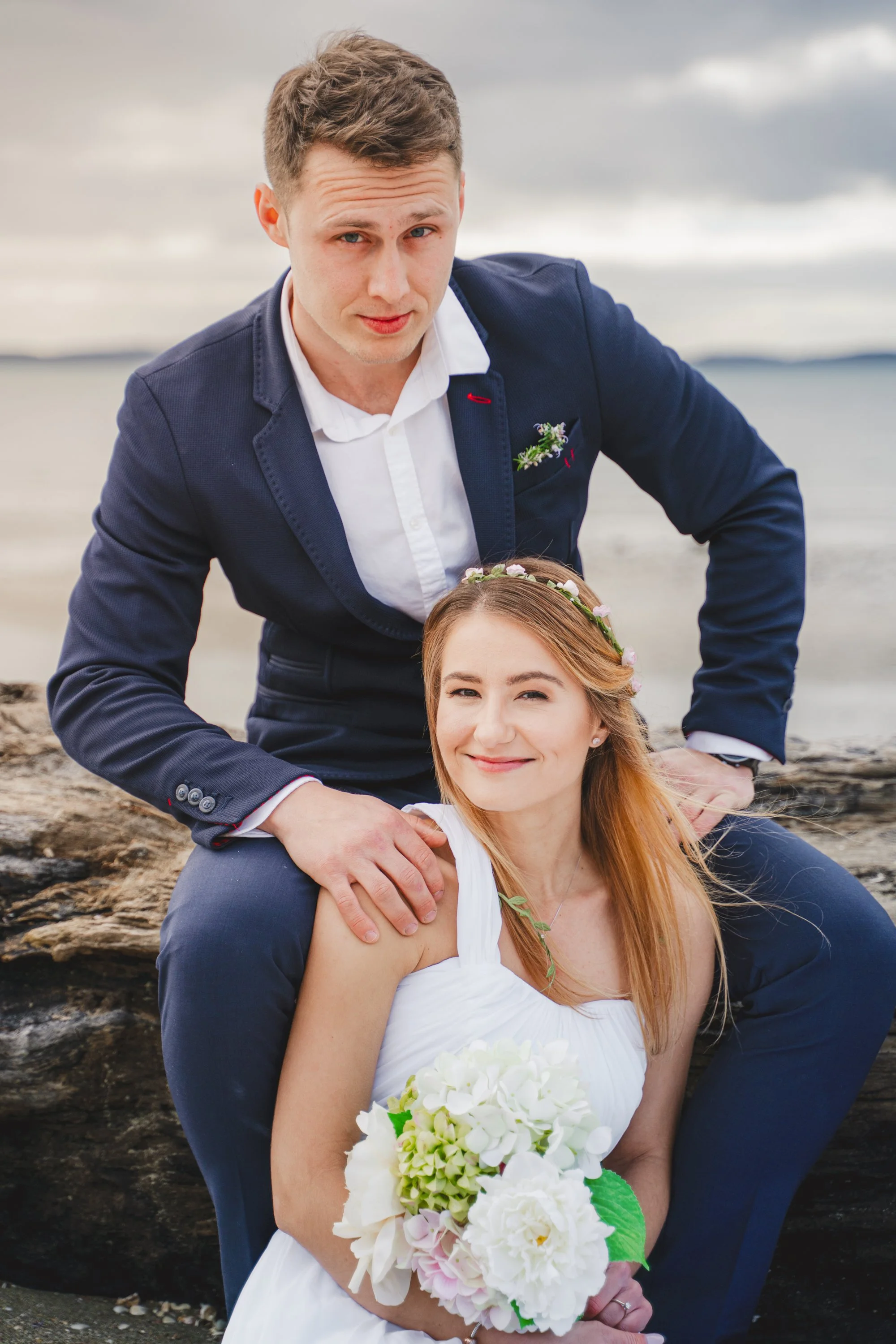 A couple dressed in wedding attire sitting on a rocky beach. The woman is holding a bouquet of white flowers and has a floral headband. The man is wearing a dark blue suit with a white shirt and has his hand on her shoulder.