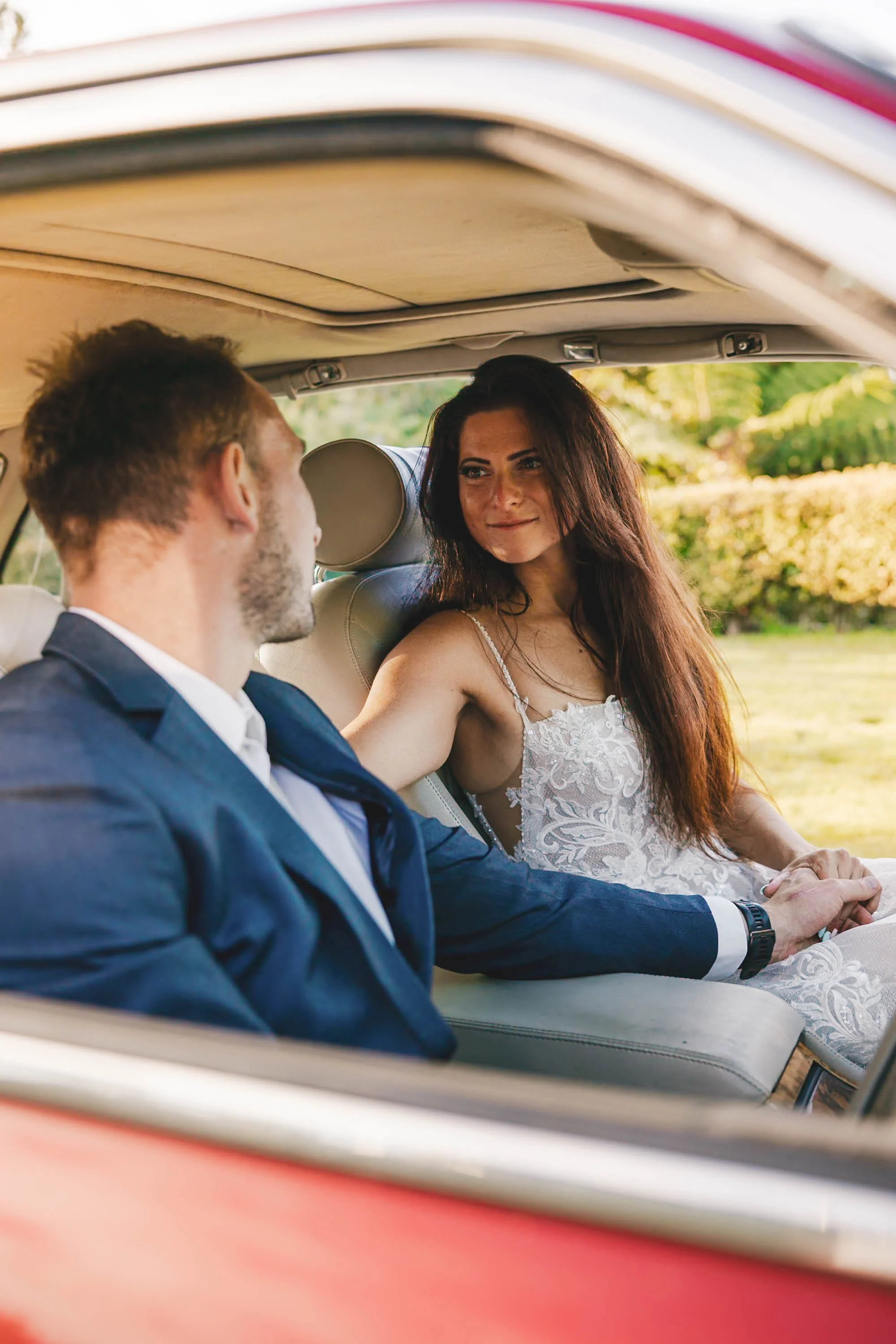 A woman in a white lace dress and a man in a navy suit sitting together in a car, holding hands, with trees and sunlight in the background.