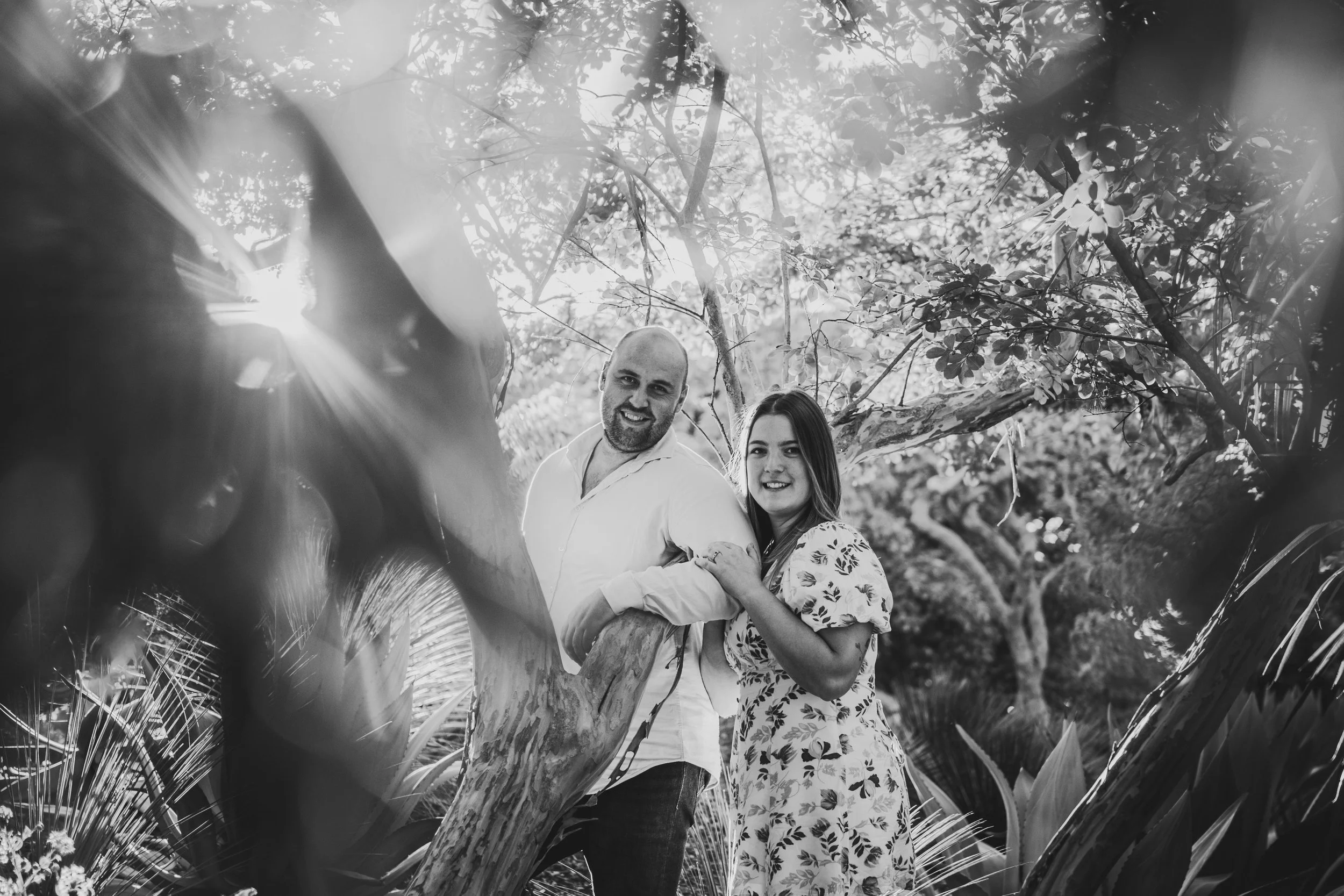 A black and white photo of a couple outdoors, standing among trees and plants, smiling at the camera.