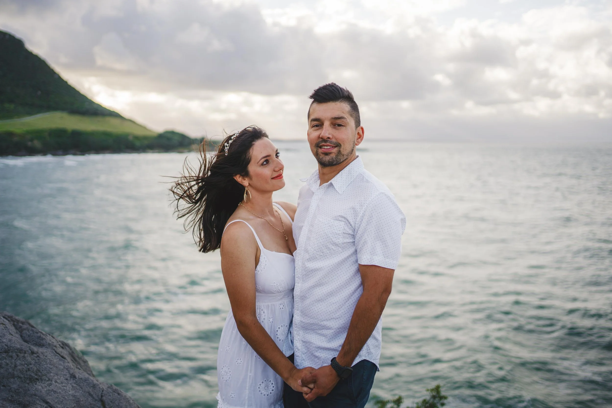 A couple standing on a rocky shoreline by the ocean, holding hands, with a green hill in the background and cloudy sky.