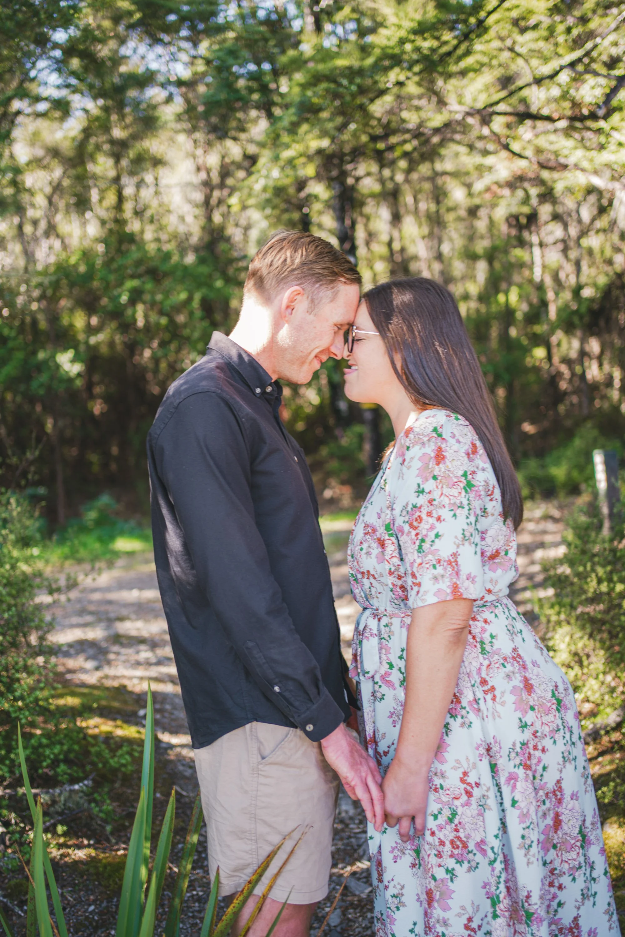 A couple stands close together in a wooded outdoor setting, touching foreheads and holding hands, both smiling.