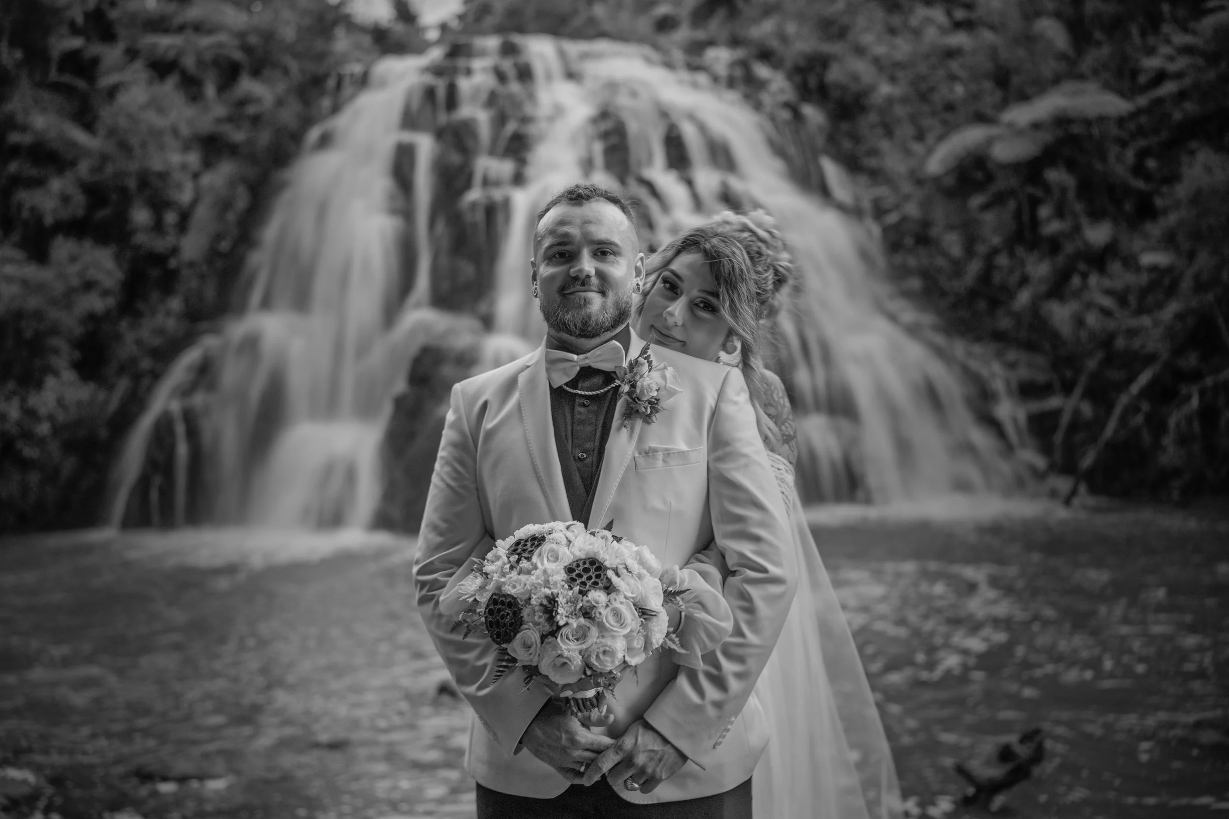 A black-and-white photo of a bride and groom standing in front of a waterfall. The groom, wearing a light-colored suit and bowtie, holds a bouquet of flowers. The bride stands behind him, leaning her head on his shoulder.