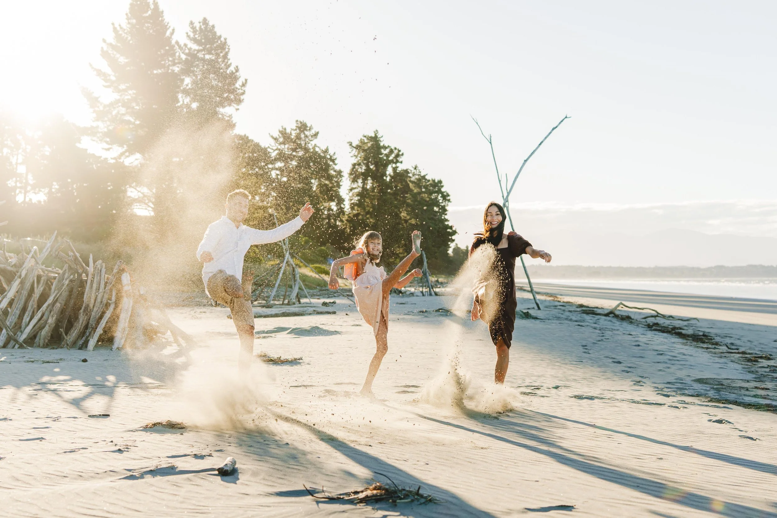 Family of four playing and kicking up sand on the beach during sunset, with trees in the background.