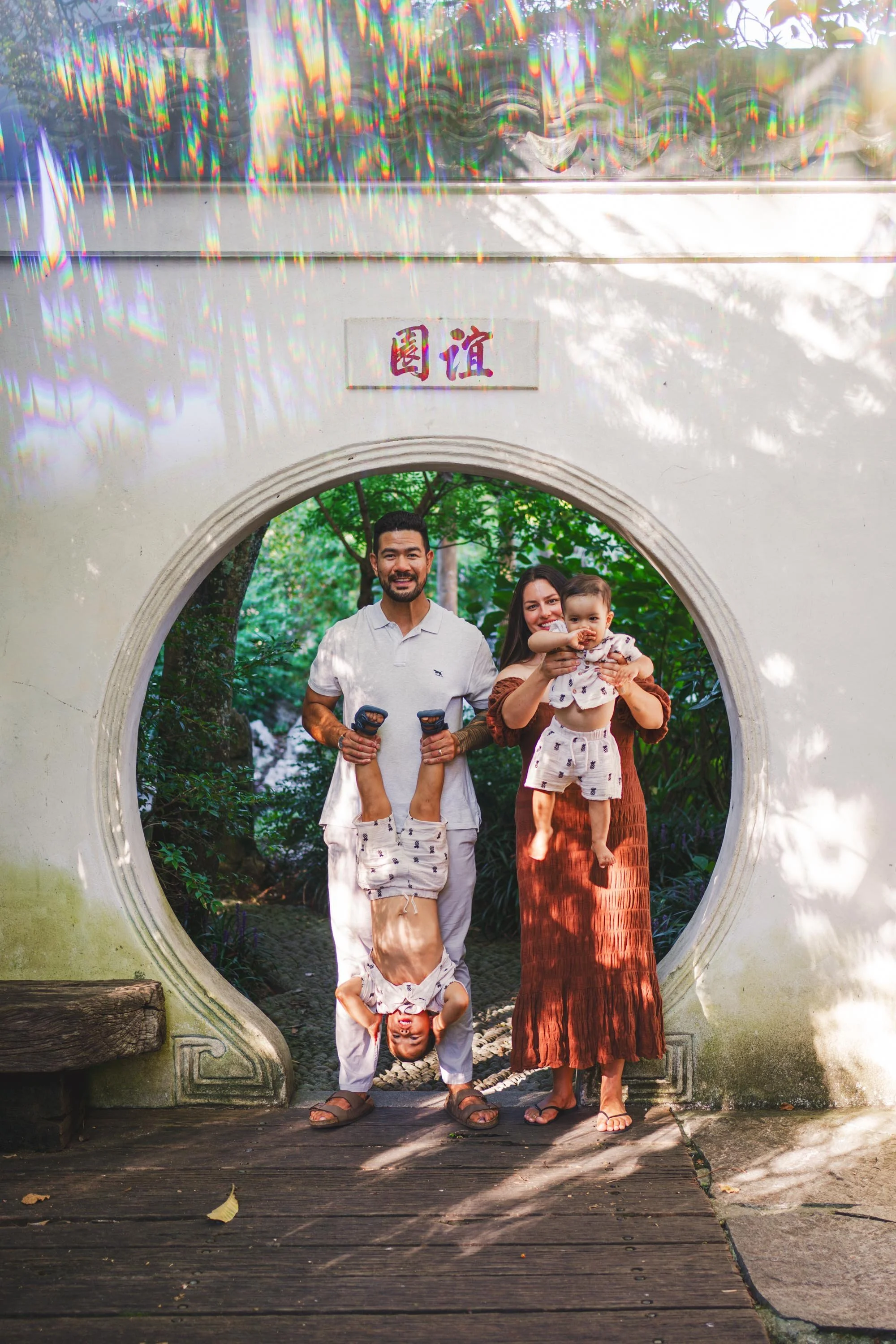 A family of four posing together in front of a traditional Chinese moon gate at a park, with trees and greenery in the background. One child is doing a handstand, another child is being held by a woman, and a man is standing beside them.