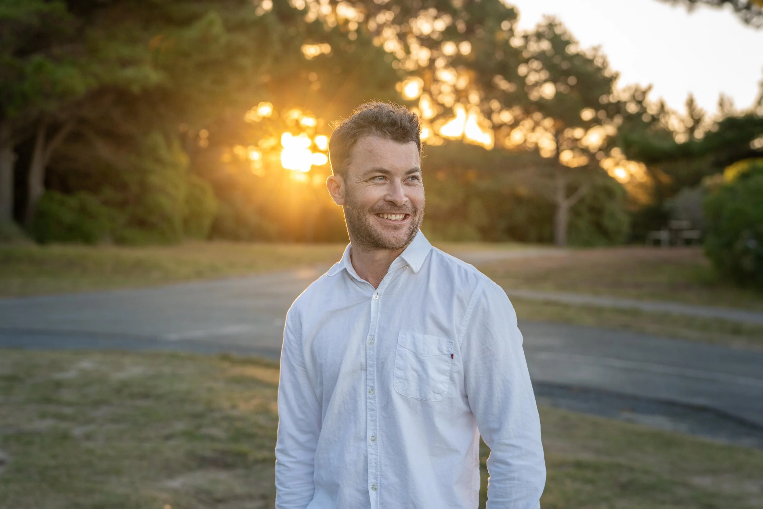 A smiling man in a white shirt standing outdoors during sunset, with trees and a path in the background.