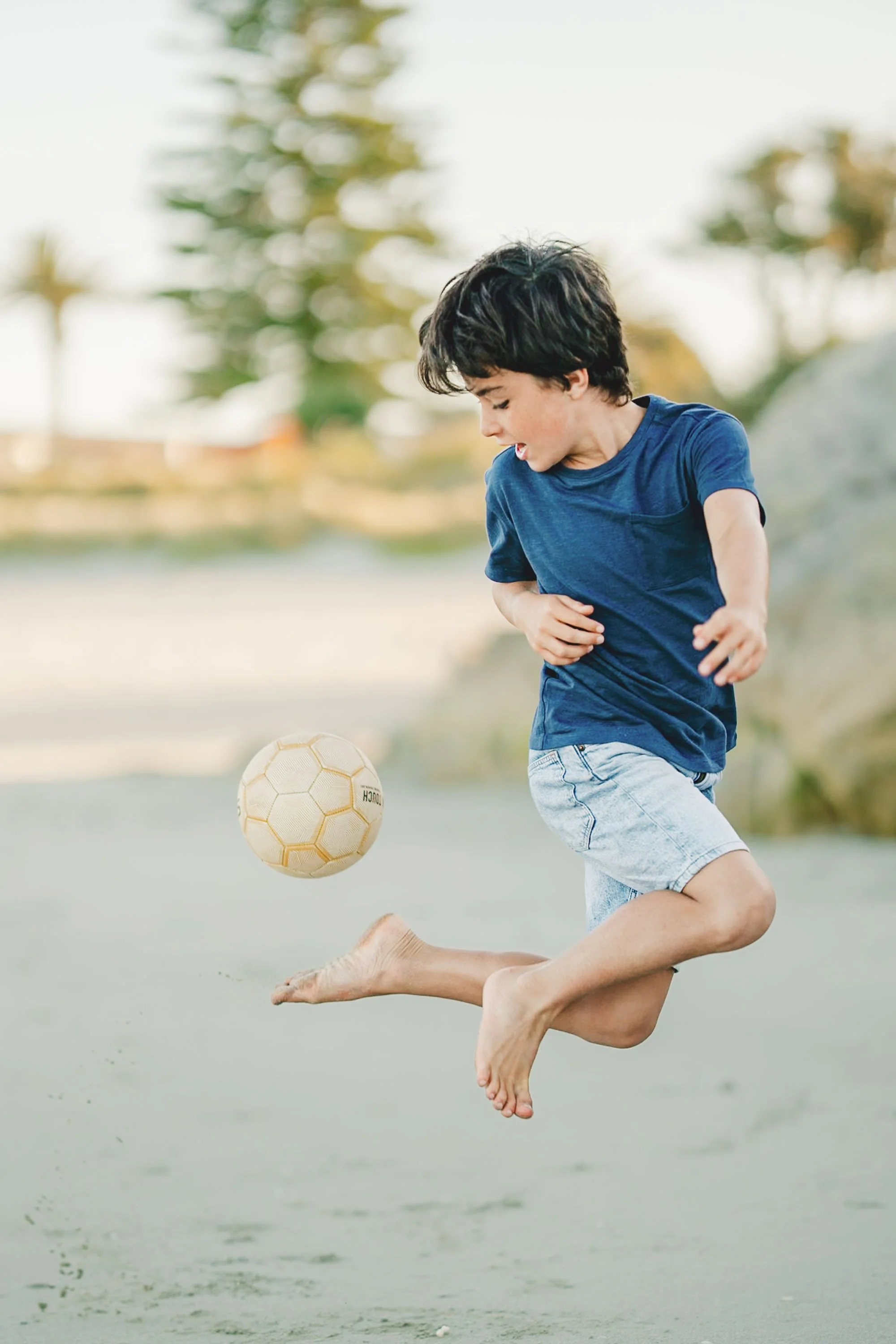 Young boy jumping barefoot on a sandy surface, playing with a soccer ball outdoors, with trees and a bright sky in the background.