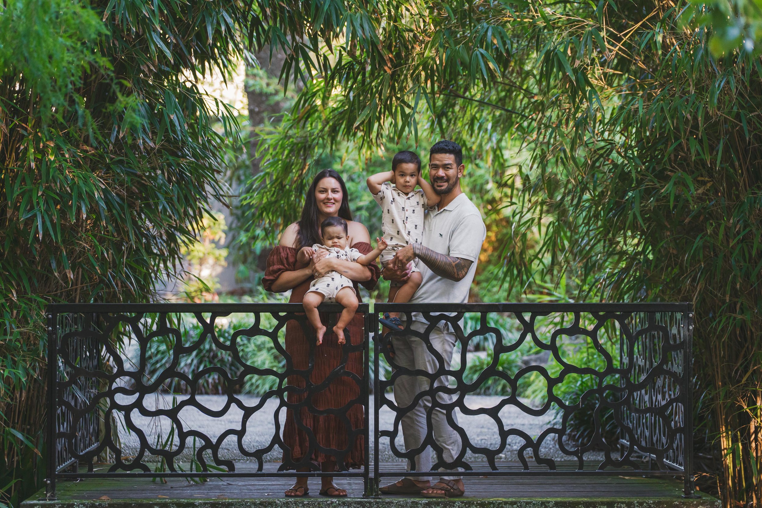 Family of four standing on a bridge with decorative railing among lush greenery, smiling and holding children in a natural outdoor setting.