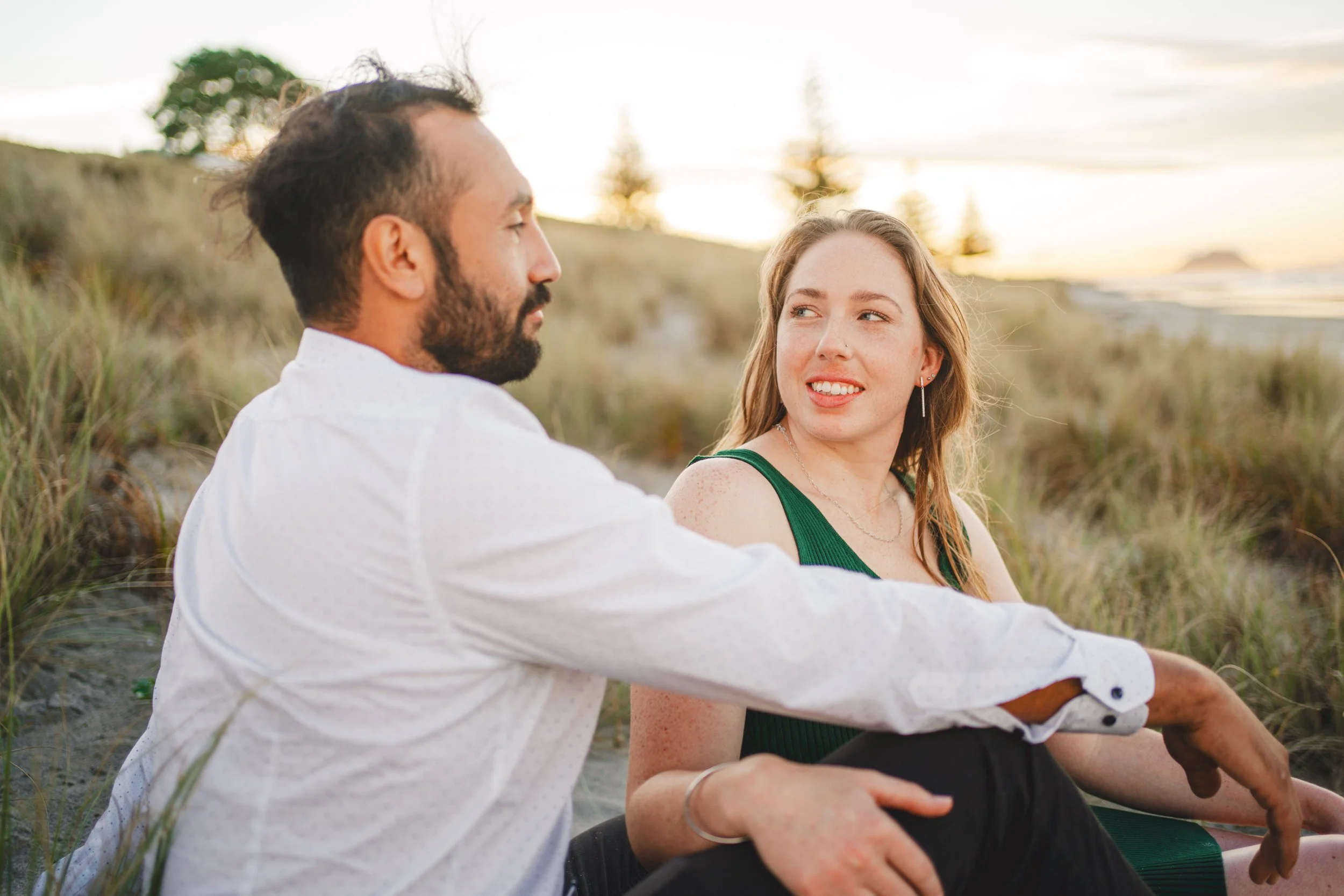 A man and a woman sitting on the beach during sunset, looking at each other with a background of dunes, trees, and the ocean.