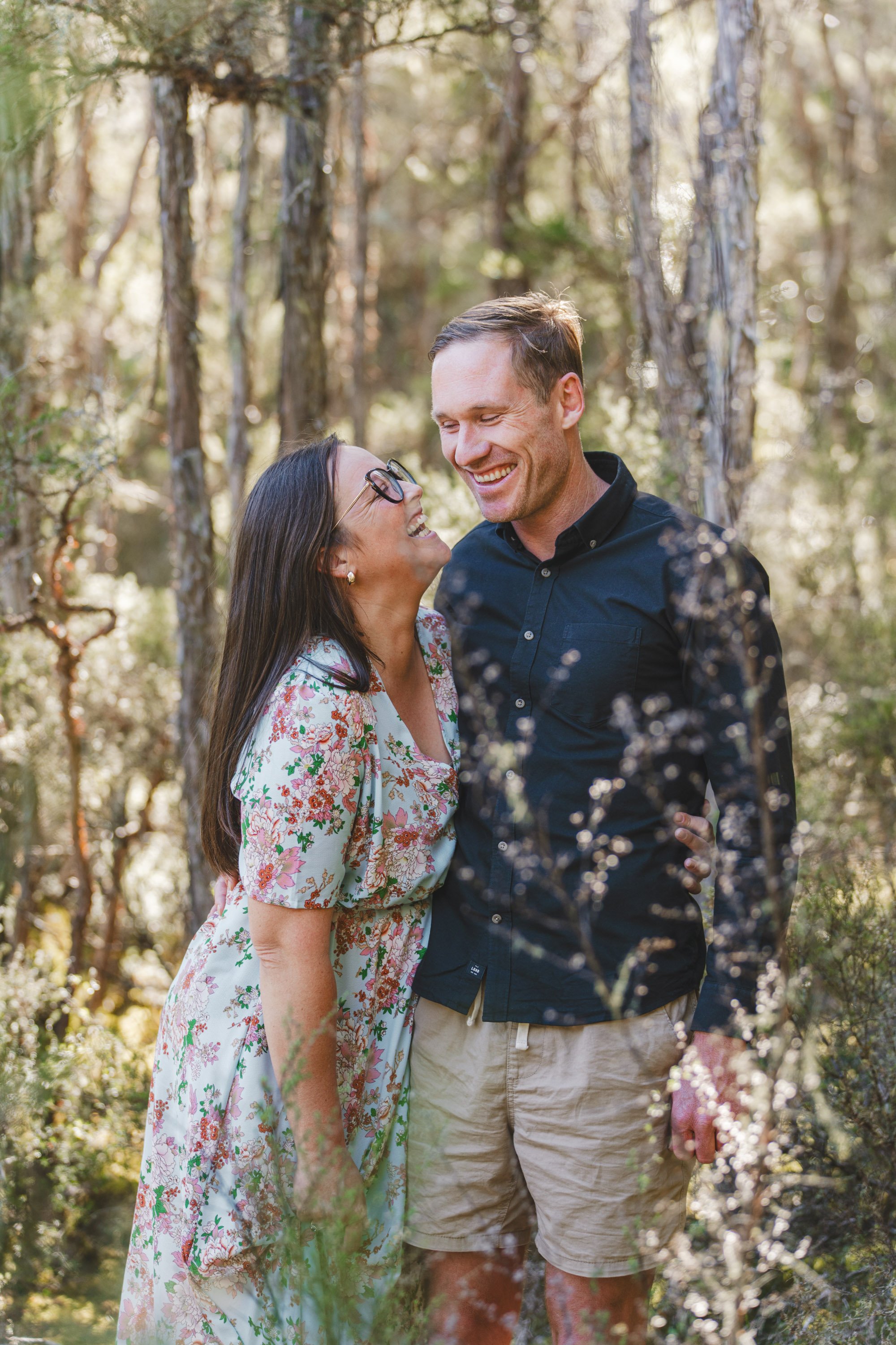 A smiling woman and man in a forest, embracing and looking at each other happily.
