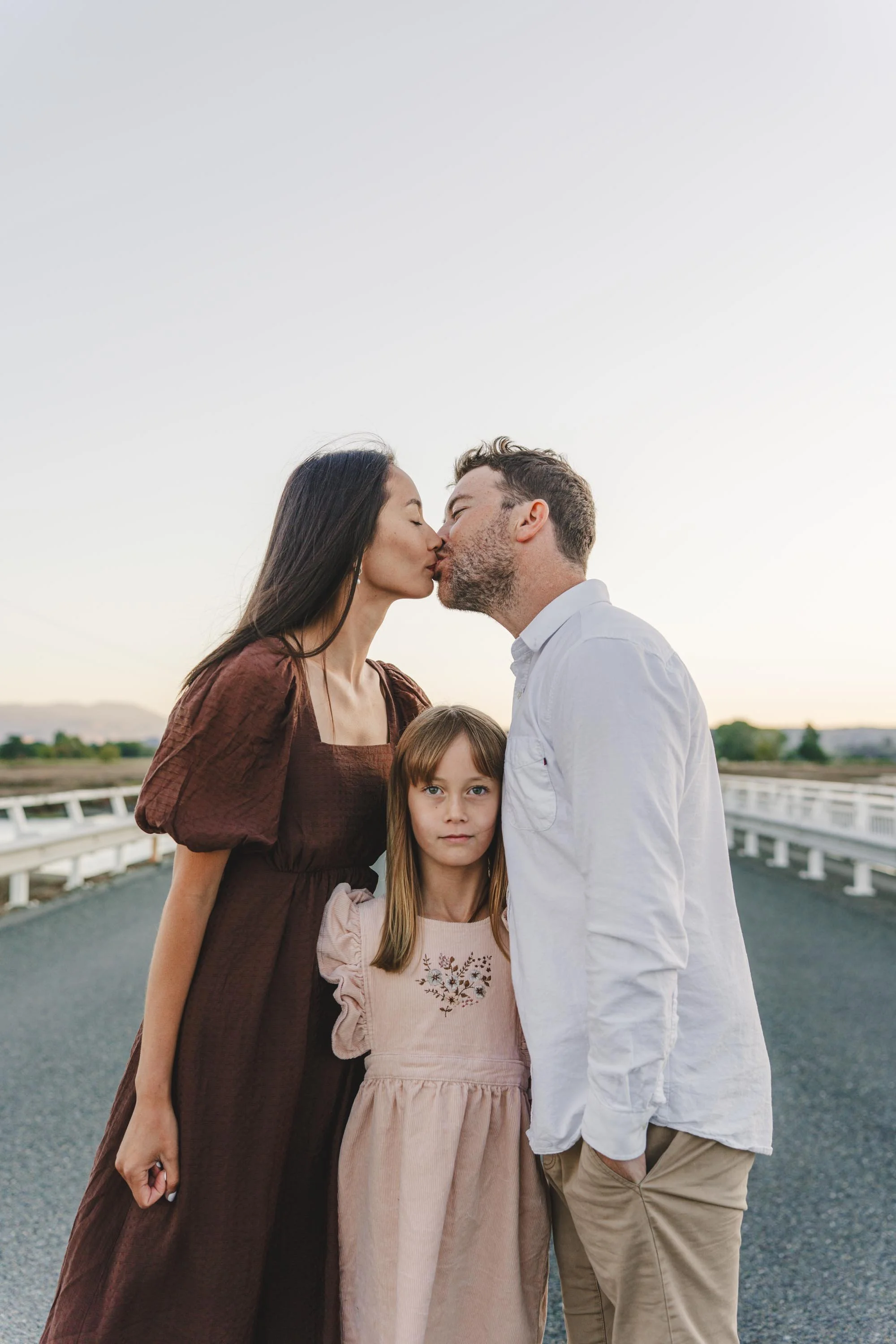 A family of three standing on an outdoor road at sunset, with the mother and father sharing a kiss and the young daughter standing in front of them looking at the camera.