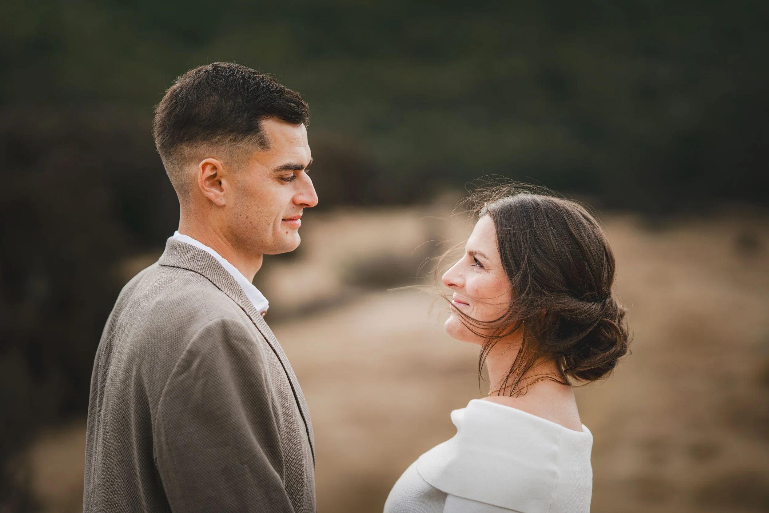 A man and woman standing face to face outdoors, gazing at each other with a blurred natural background.