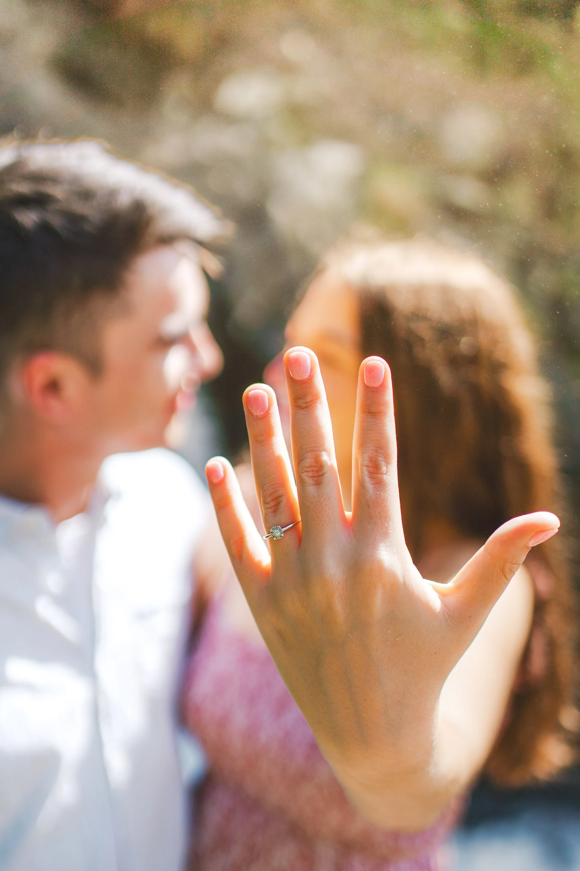 Close-up of a woman's hand showing an engagement ring, with a blurred couple in the background.