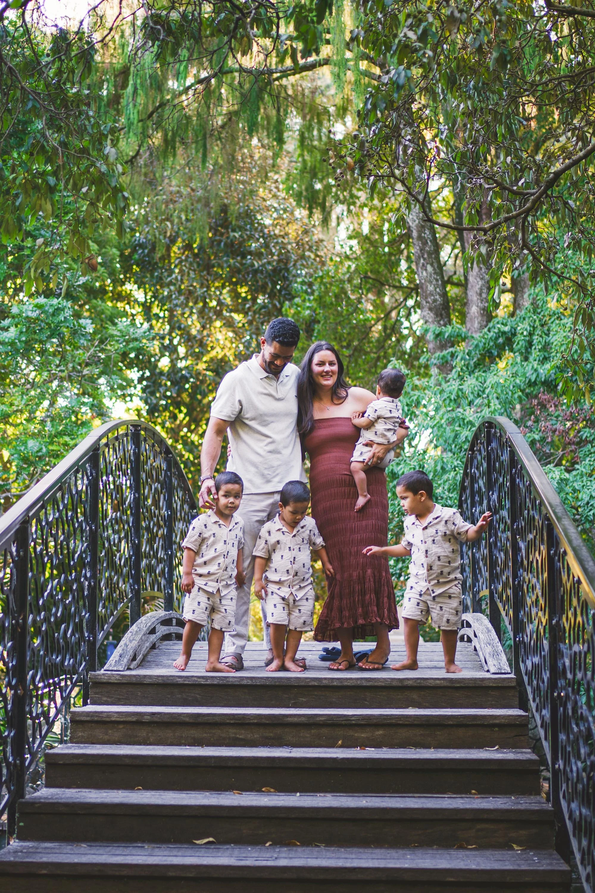 Family of six walking down a wooden bridge surrounded by lush green trees, with the mother holding a baby and all family members smiling.