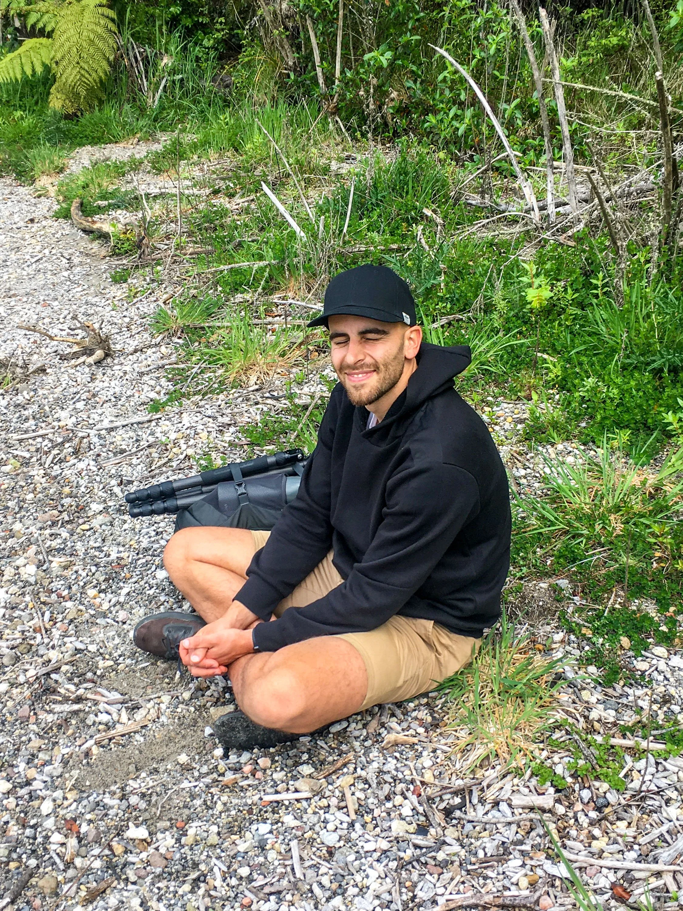 Young man sitting barefoot on a gravel trail, smiling with eyes closed, surrounded by green foliage and bushes.