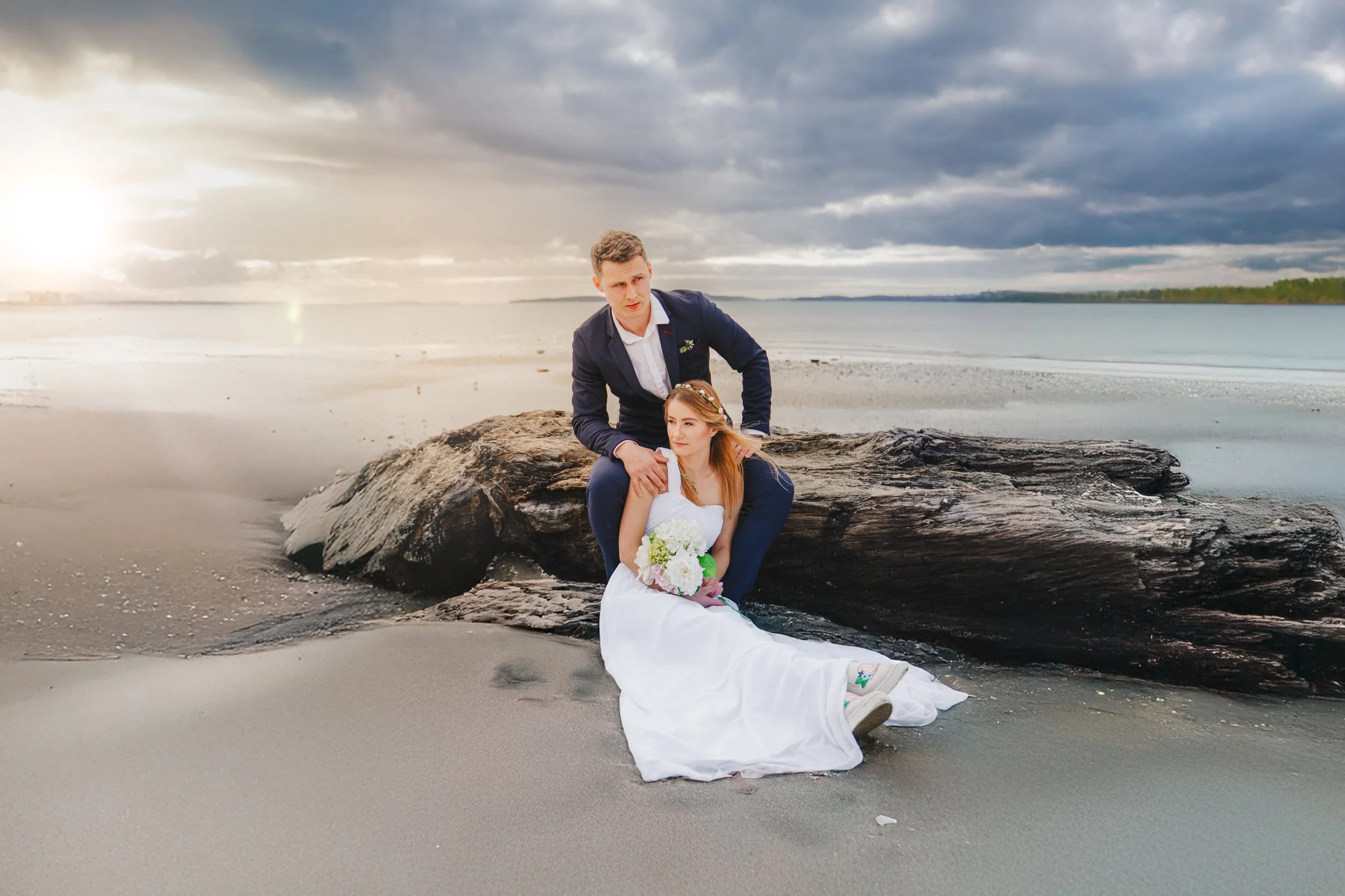 A bride in a white wedding dress holding a bouquet of white and green flowers, sitting on the sand at the beach with a groom in a dark suit kneeling beside her on a large driftwood log during sunset or sunrise.