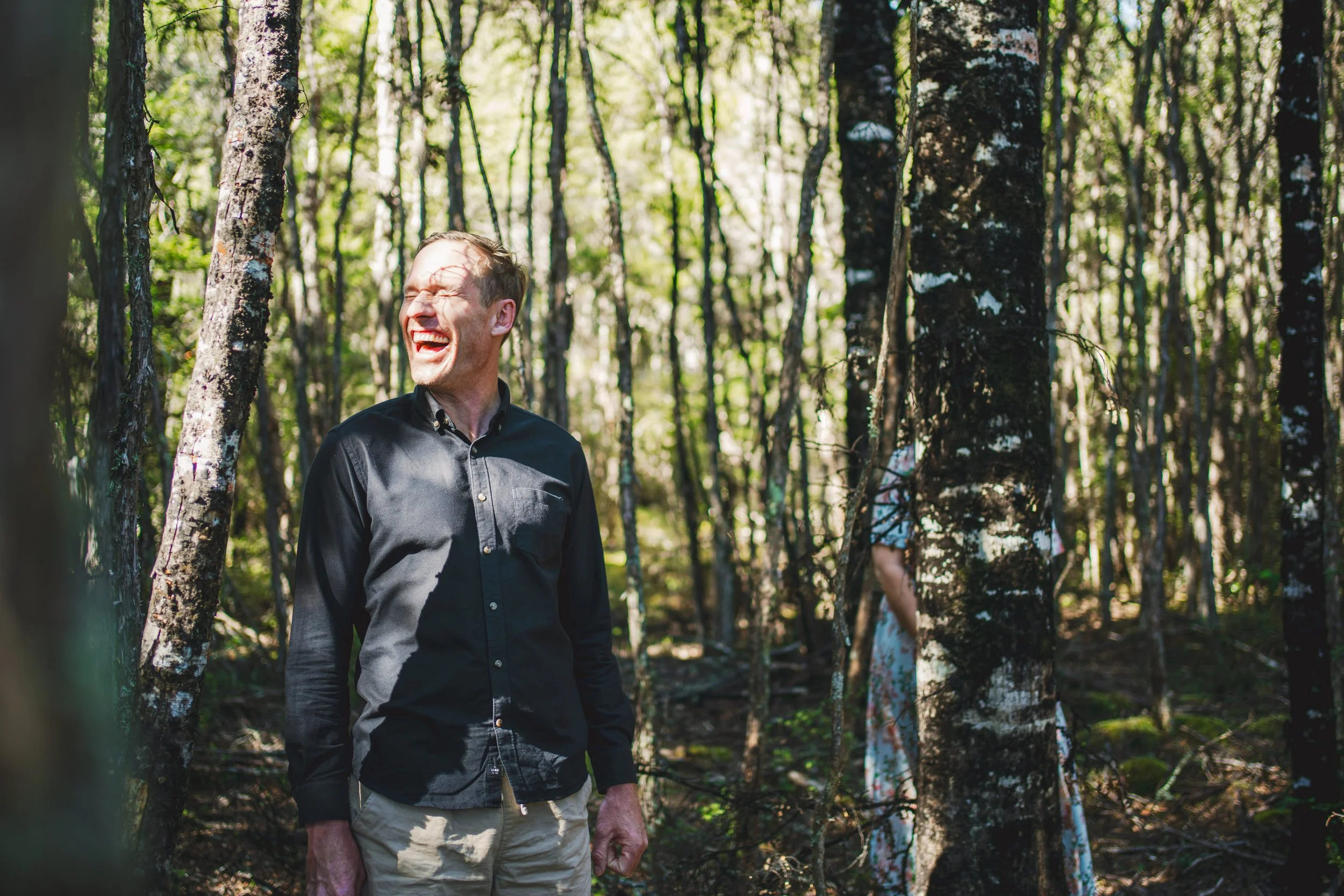 A man with light skin and short hair, wearing a black button-up shirt and tan pants, laughing in a forest with sunlight filtering through the trees.