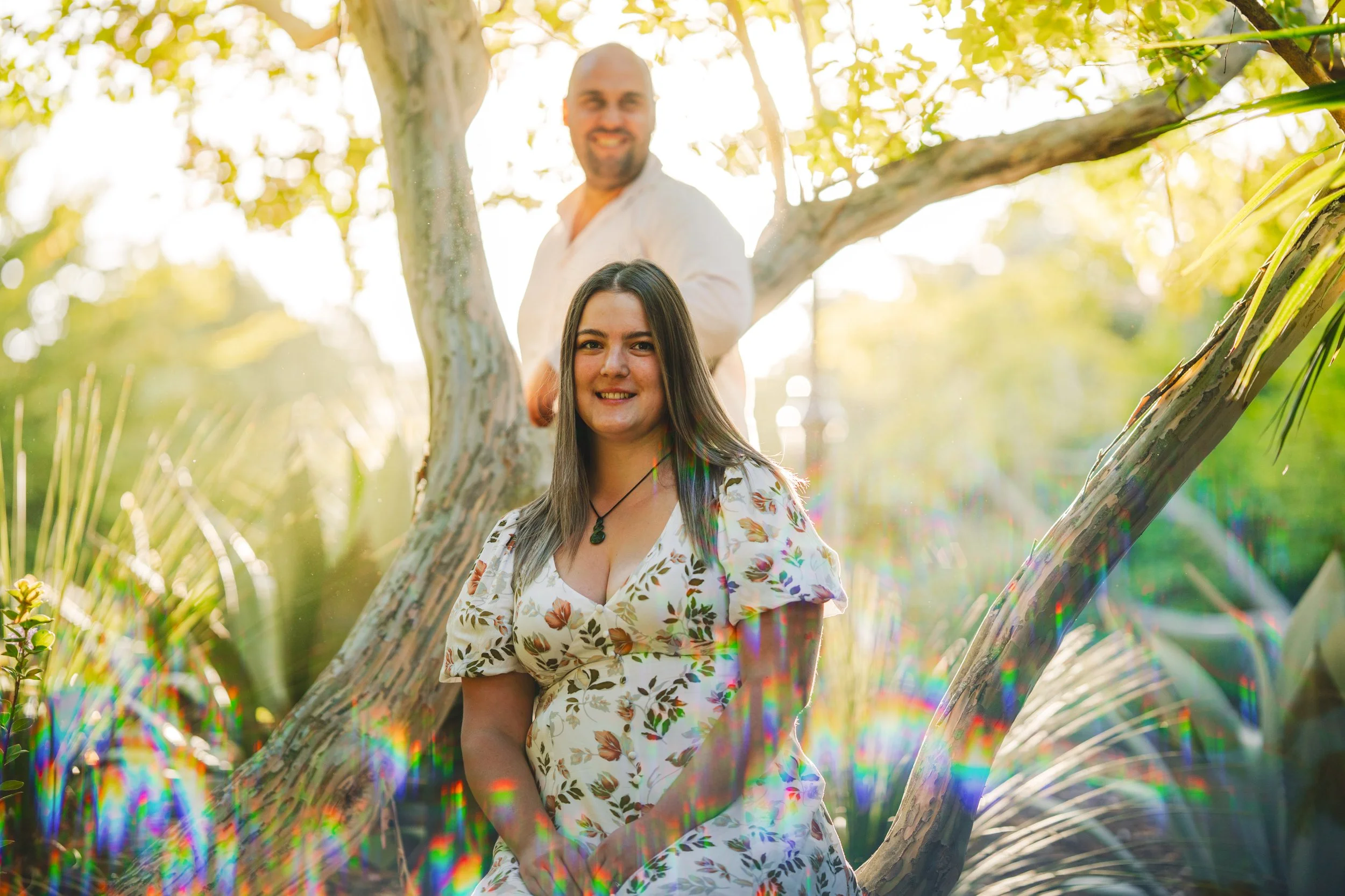 A young woman sitting on a tree branch outdoors, smiling at the camera, with a man standing behind her on the same tree, smiling at the camera, surrounded by greenery and sunlight.