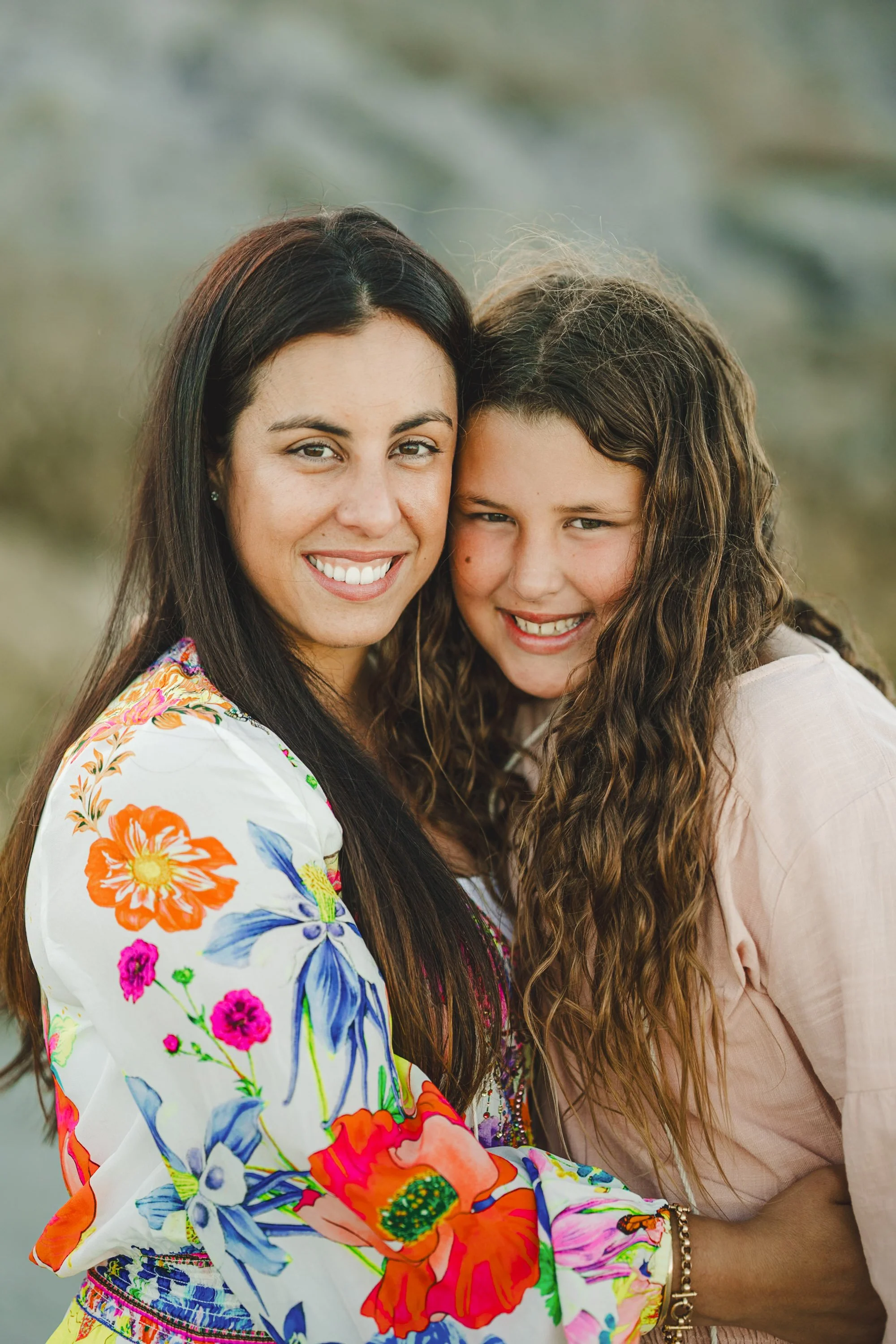 A woman and a girl smiling and hugging outdoors, woman wearing a colorful floral dress and girl in a light-colored shirt.