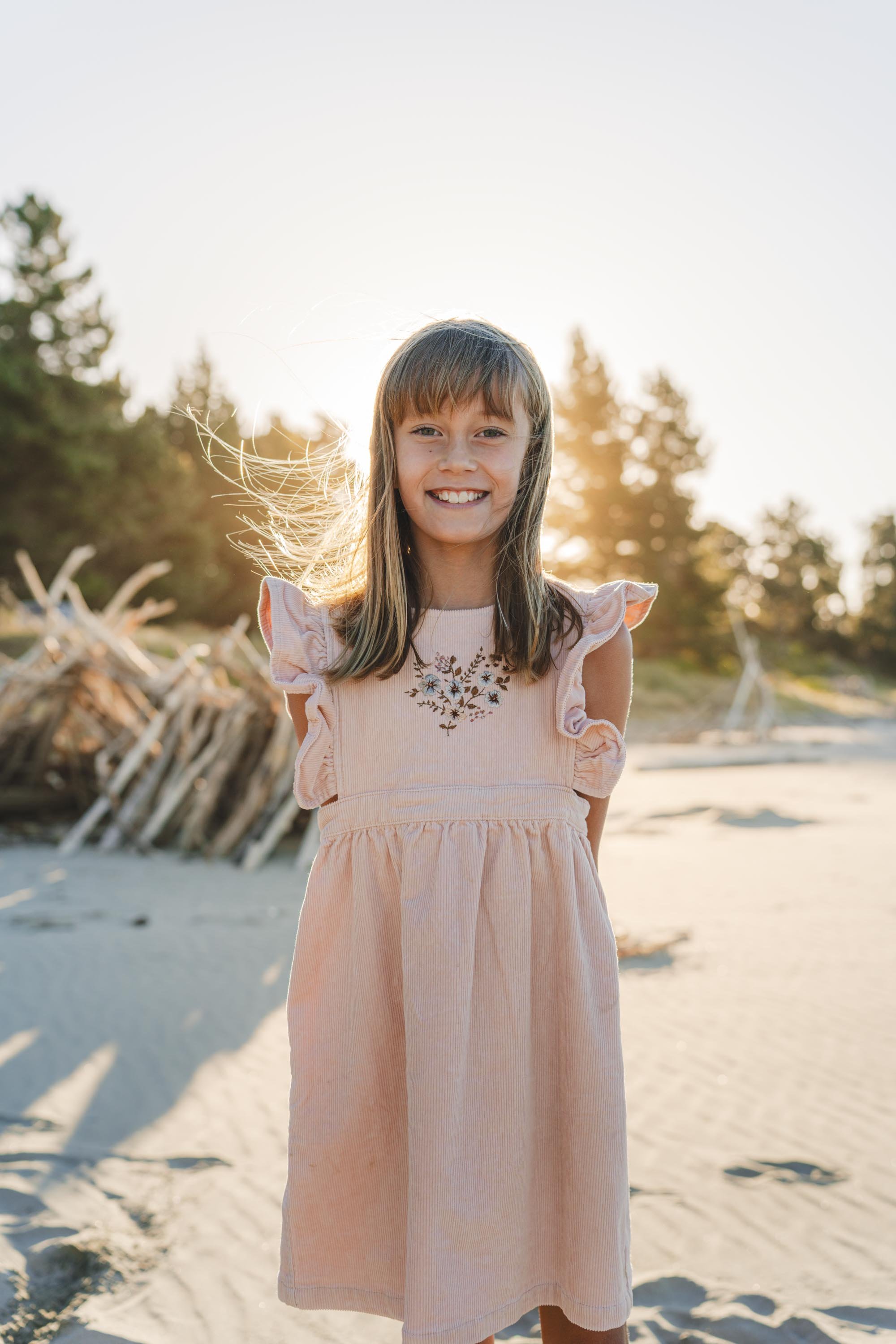 A smiling young girl in a pink dress standing on a sandy beach with trees and driftwood in the background, backlit by the setting sun.