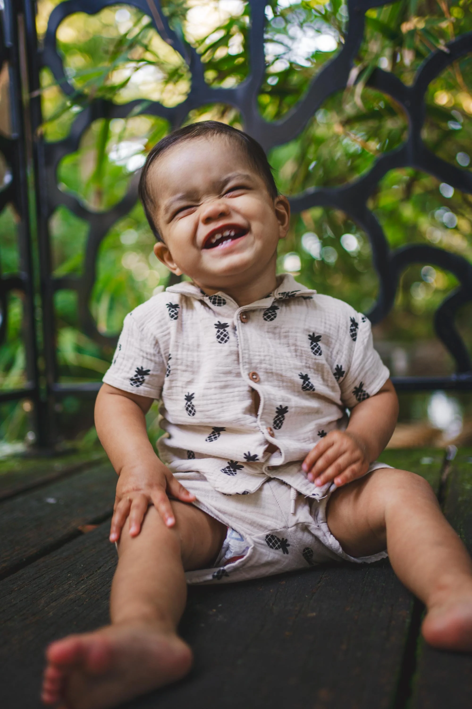 A young child sitting outdoors on a wooden surface, smiling with eyes closed, wearing a beige shirt with pineapple patterns.