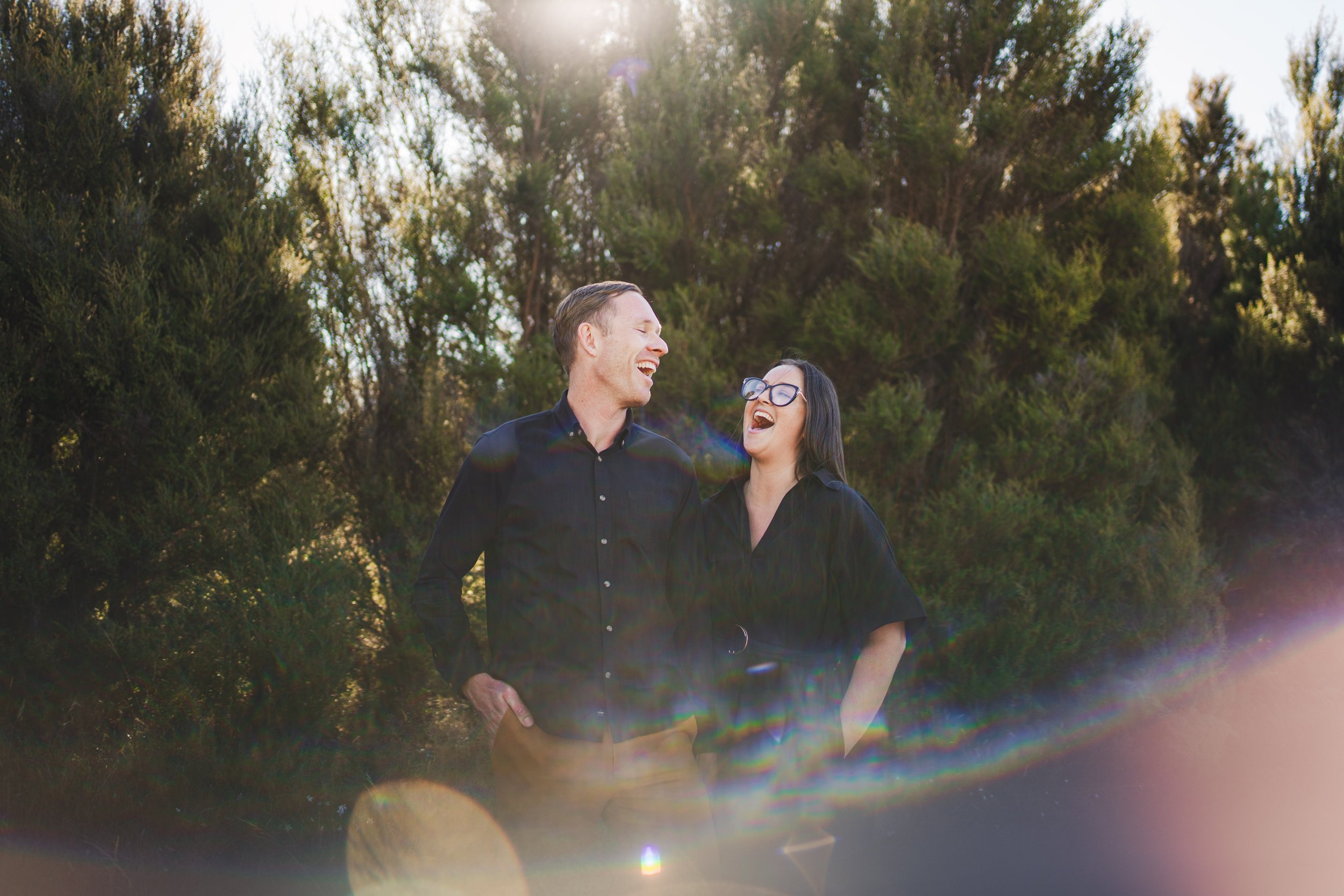 A man and woman standing outdoors, laughing and enjoying each other's company during a sunny day. The background features trees and sunlight.