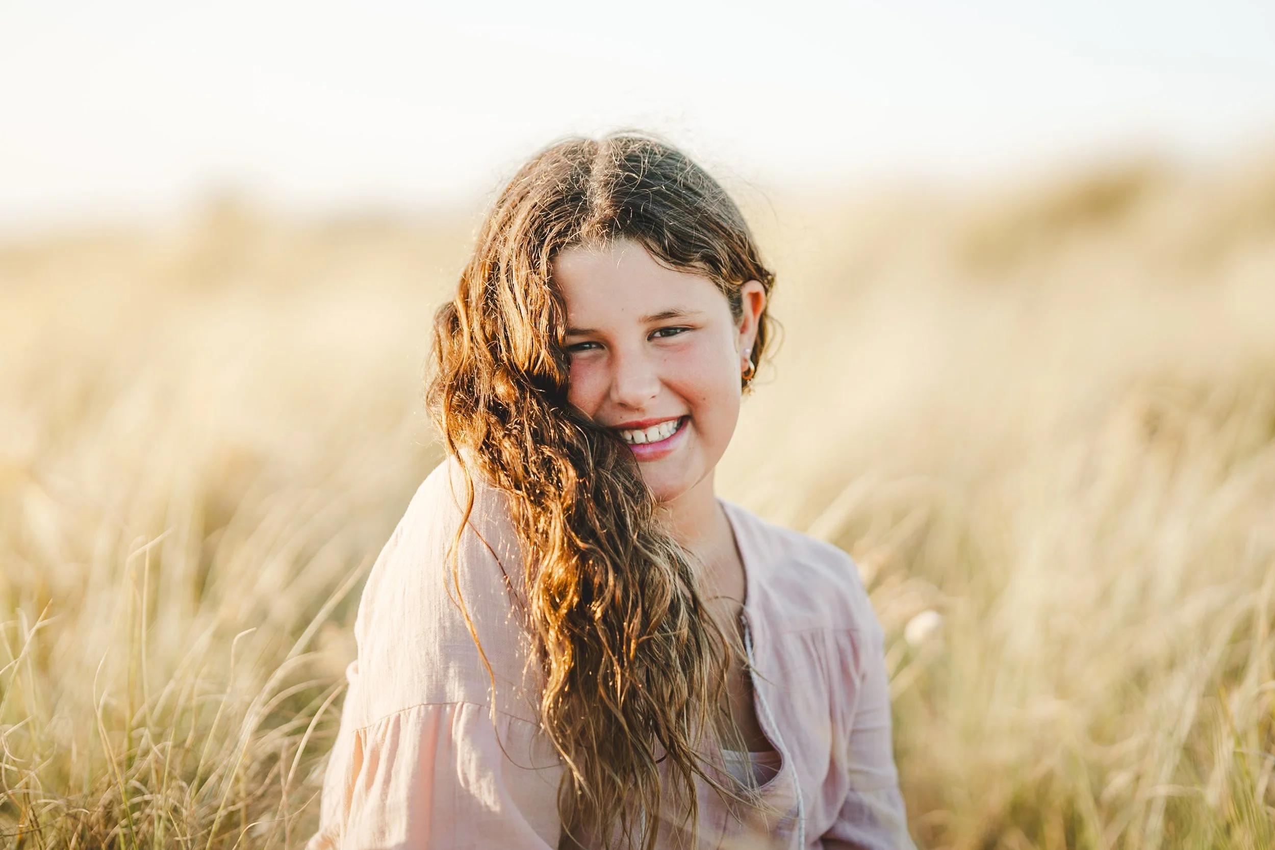 A girl with curly brown hair smiling outdoors in a grassy field during golden hour.