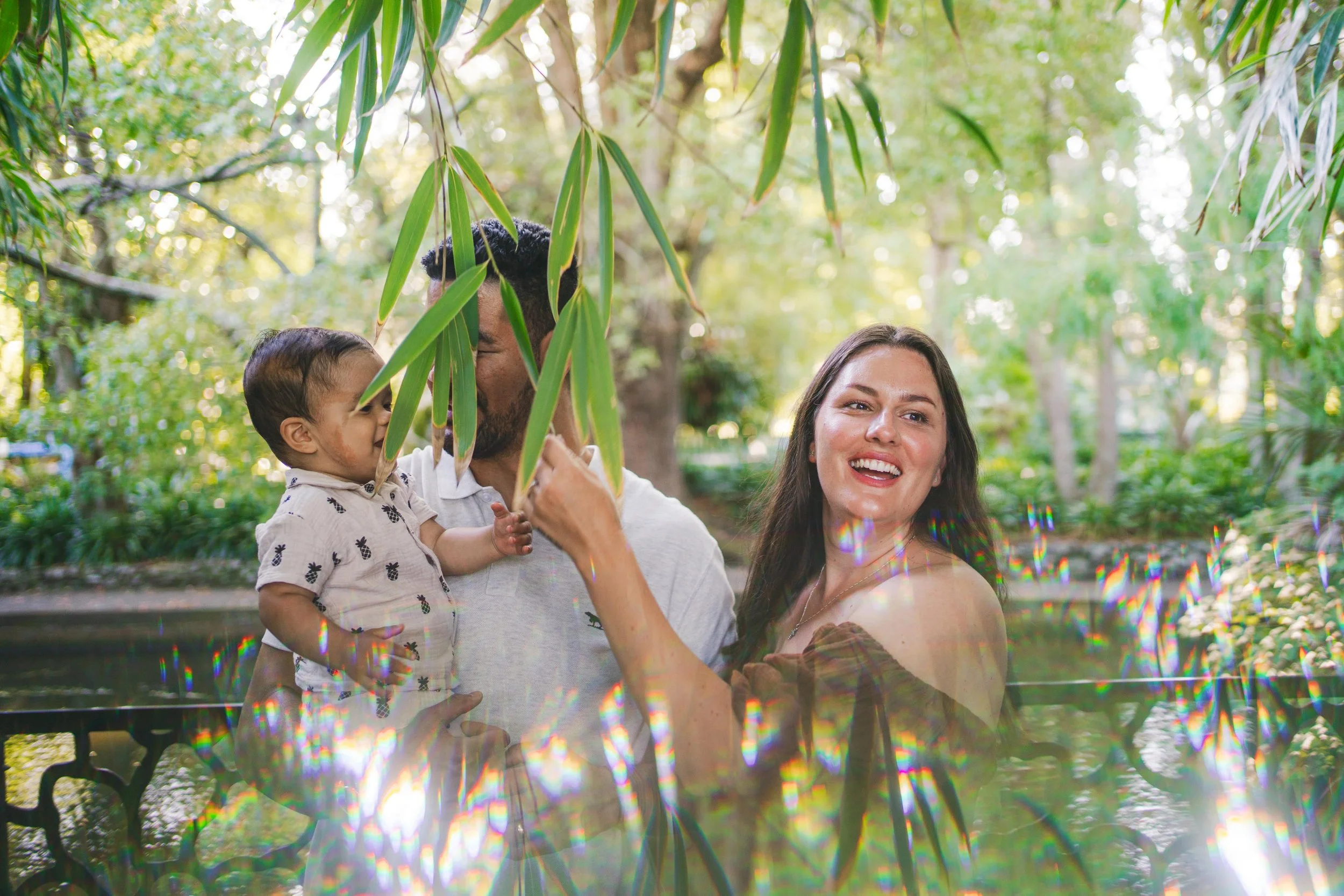 A family of three enjoying a day in a lush, green outdoor setting during daytime, with the mother smiling at the camera, the father holding a young child, and surrounded by greenery and dappled light.