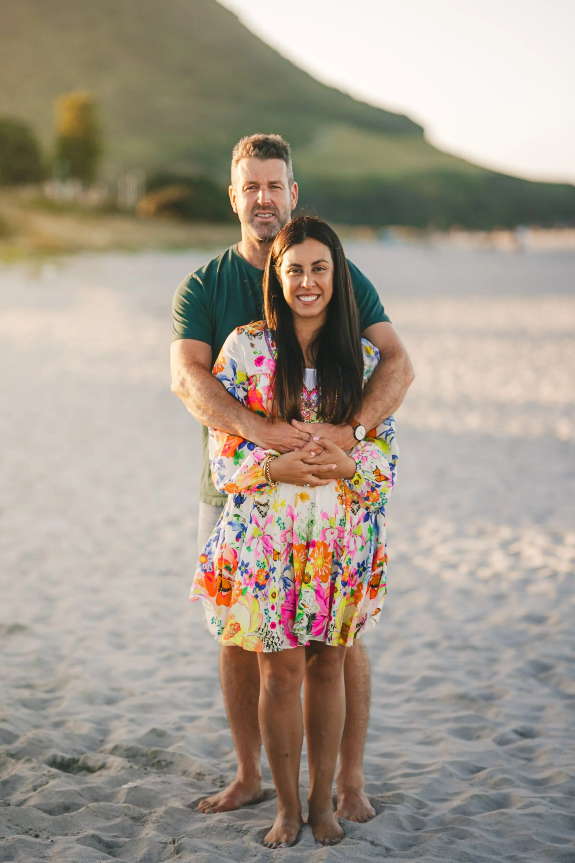 A couple standing on the beach with the man embracing the woman from behind. The woman is smiling and wearing a colorful floral dress, while the man is looking at the camera. There are hills and water in the background, and the scene appears to be du