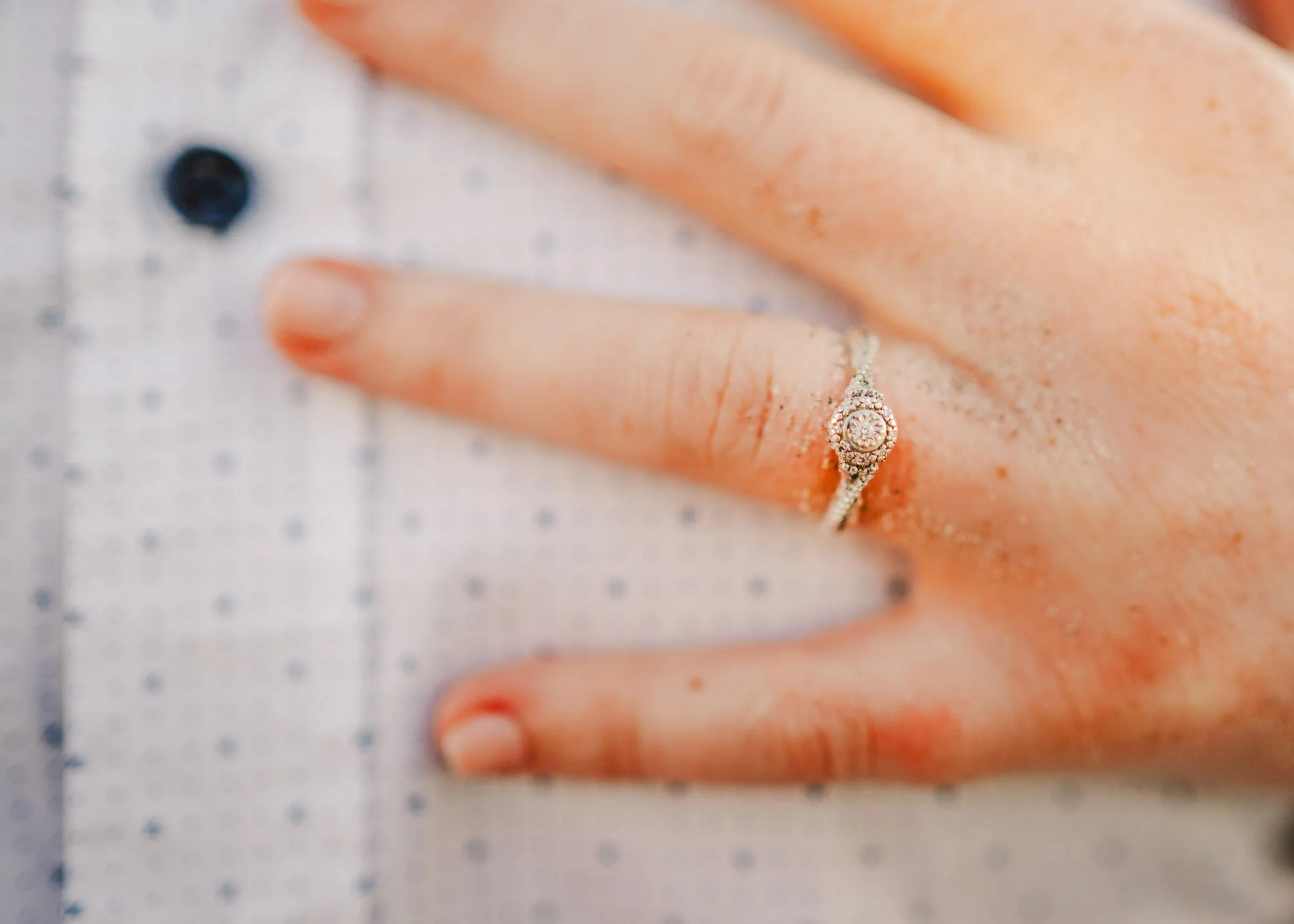 Close-up of a hand with glittery sand on fingers and an engagement ring with a round diamond in a halo setting on the ring finger.