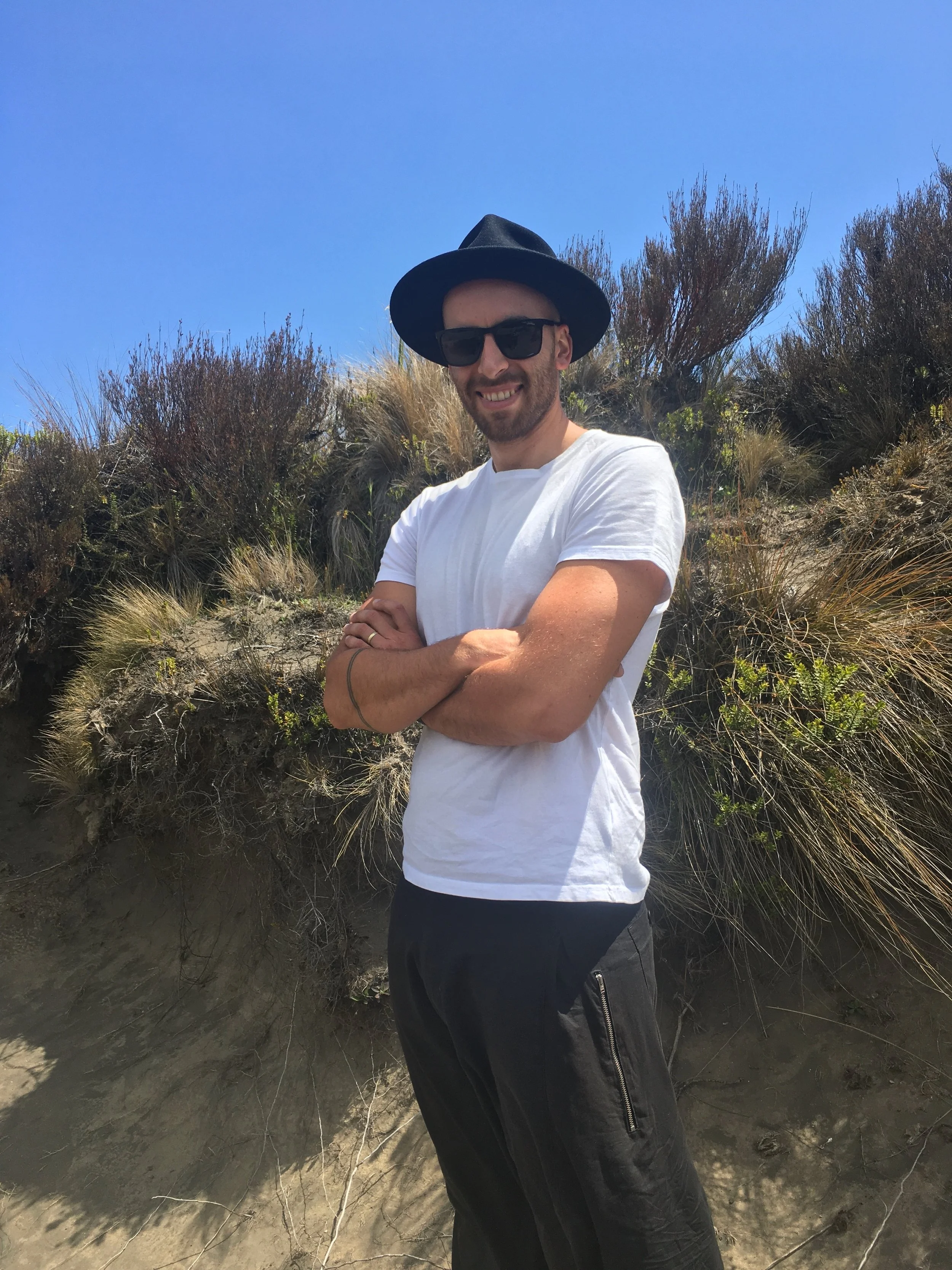 A man smiling outdoors, wearing sunglasses, a black wide-brimmed hat, a white T-shirt, and black pants, standing on a sandy hillside with dry plants and bushes under a clear blue sky.