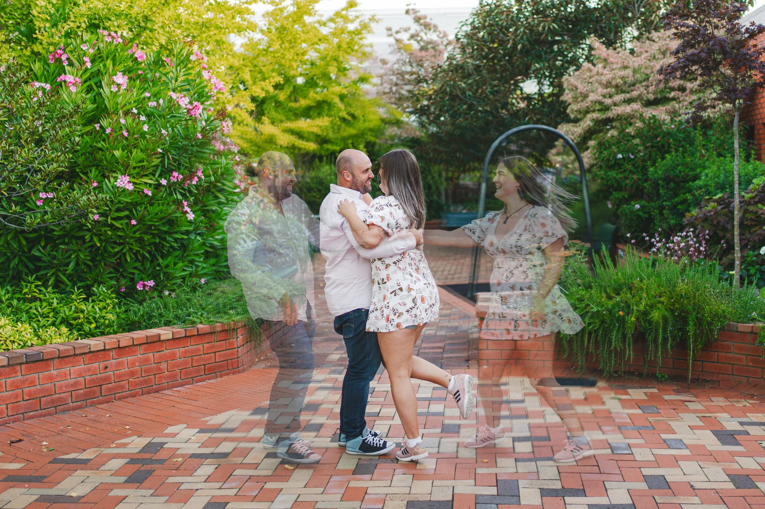 A couple dancing in a brick-paved garden, with a transparent overlay showing their past and future selves. The garden has lush greenery, pink flowers, and trees, with a peaceful background.