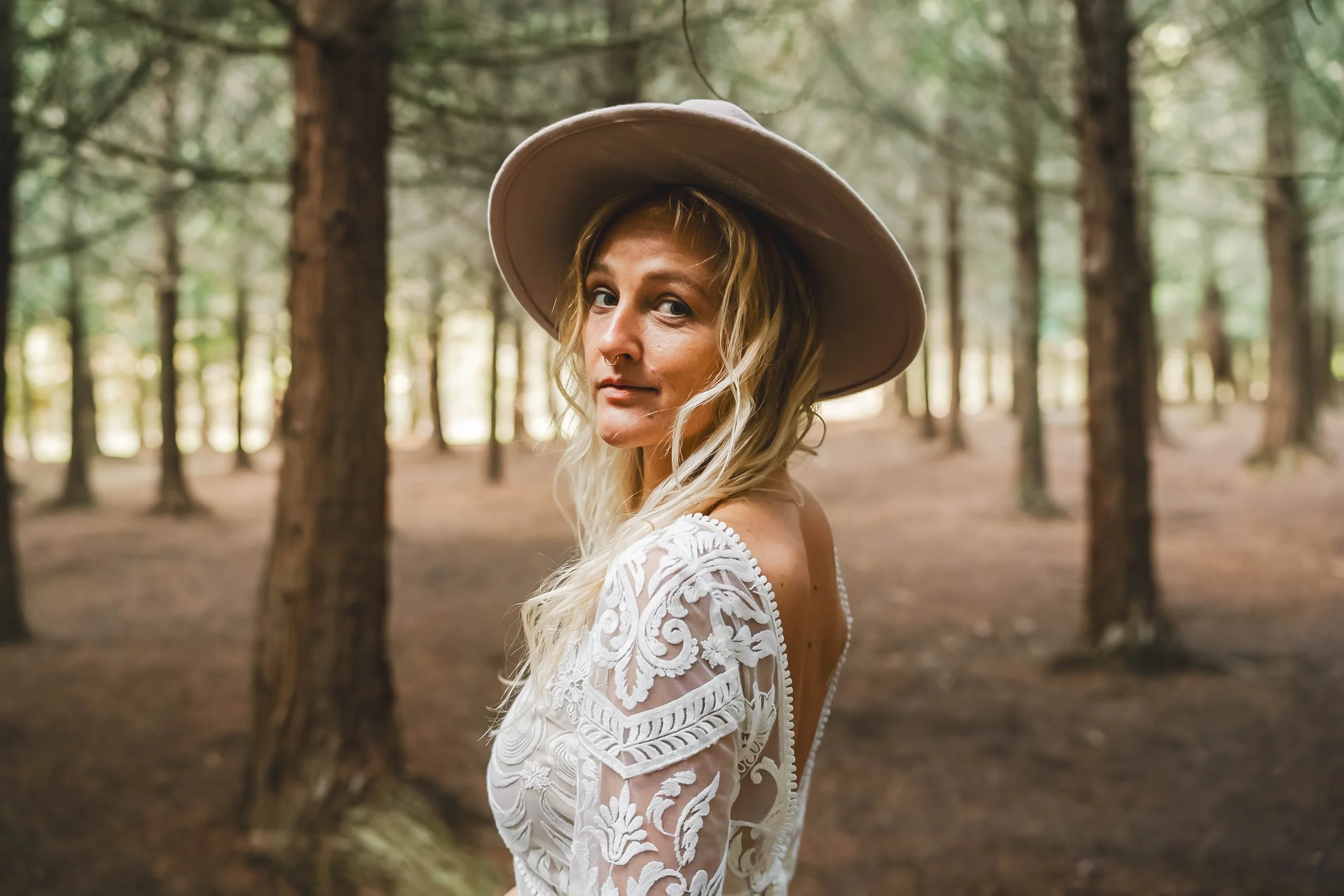 A woman with long blonde hair wearing a large beige hat and a white lace dress standing in a forest with tall trees.