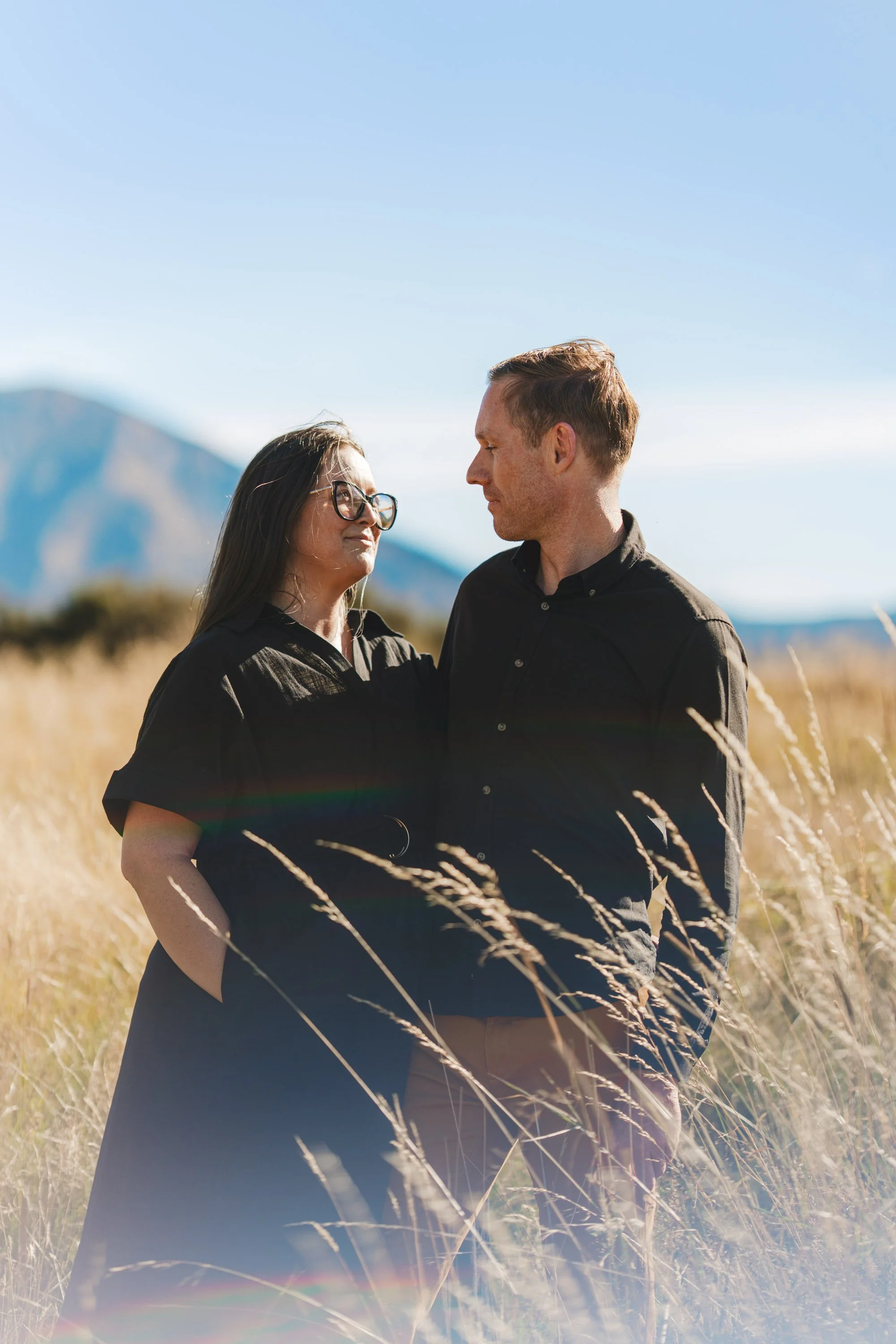 A man and a woman standing close together in a field of tall grass with mountains in the background, looking at each other.