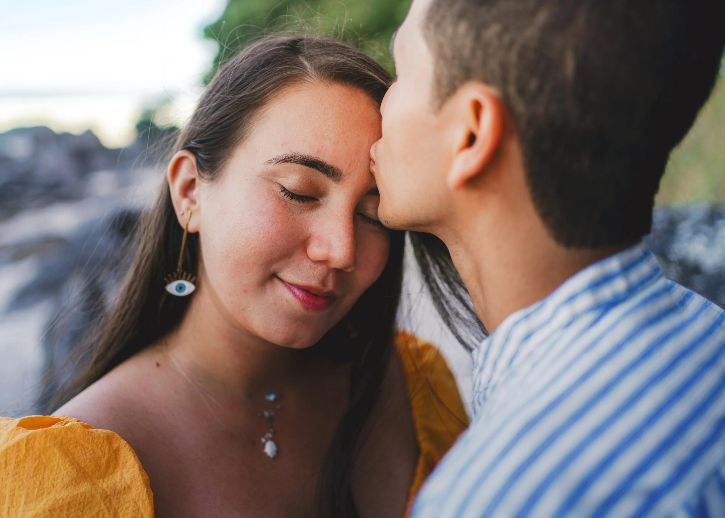A woman with long dark hair and a man with short dark hair sharing a tender moment with foreheads touching outdoors.