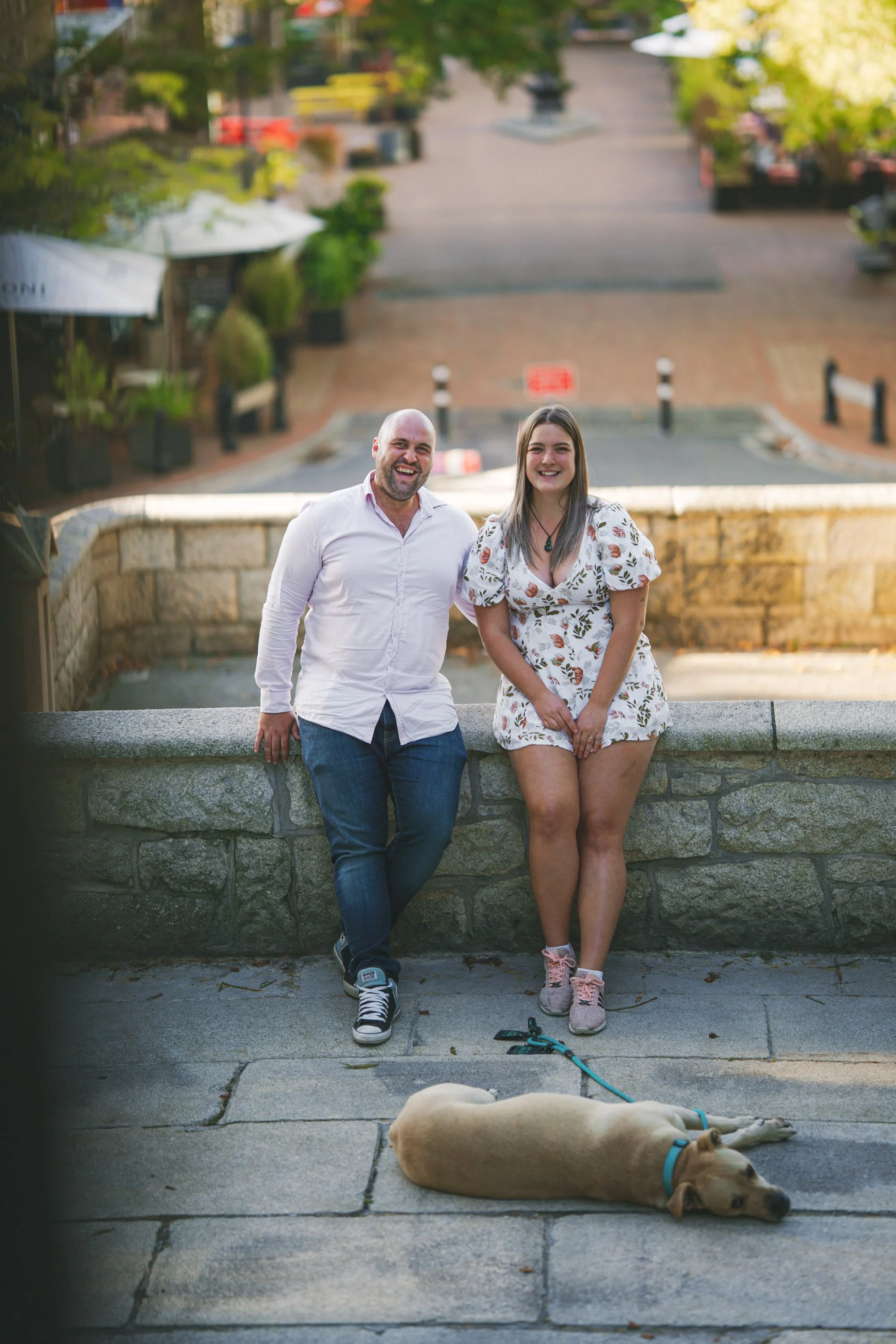 A man and woman are smiling and standing against a stone wall outdoors, with a dog lying on the ground in front of them, in a park or town square setting with trees and brick path behind them.