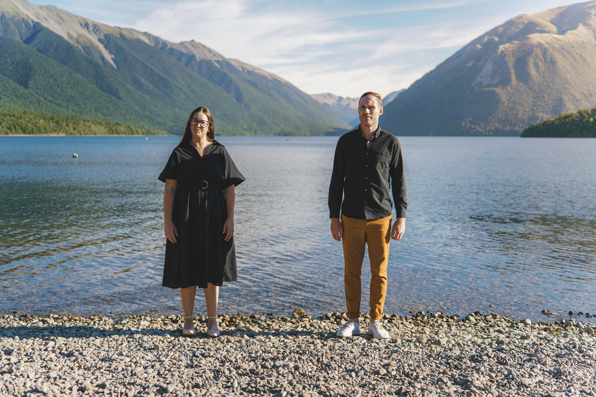 A man and woman standing on a pebbled beach in front of a lake surrounded by green mountains on a clear day.