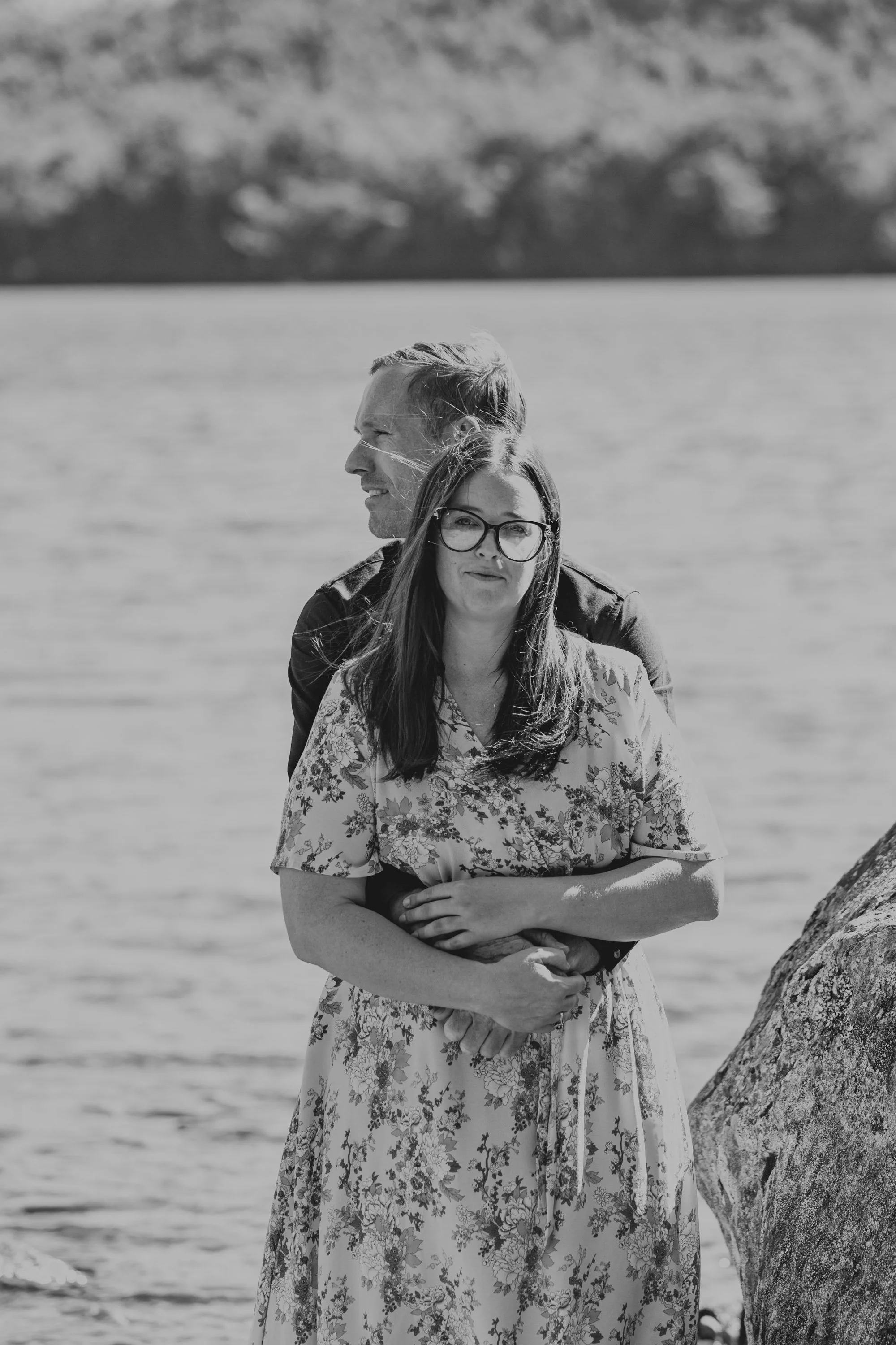 A black and white photo of a couple standing by a lake; the woman is wearing glasses and a floral dress, and the man is embracing her from behind, both looking at the camera.