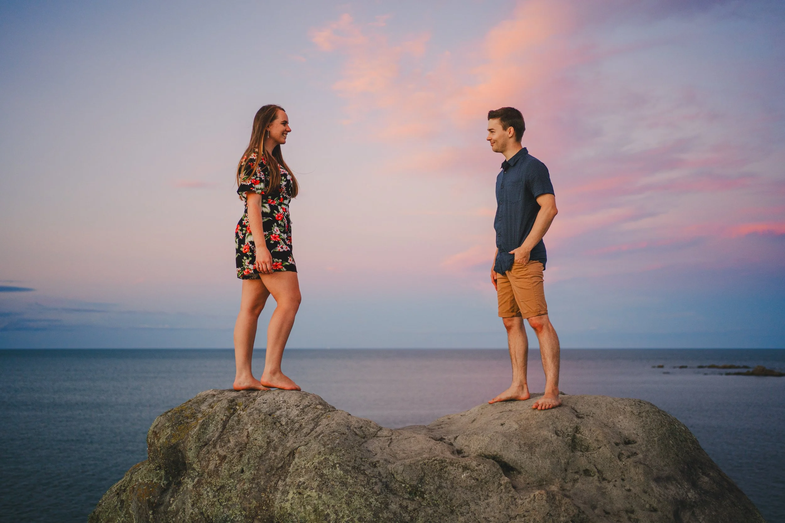 A man and woman standing on separate rocks near the ocean during sunset, facing each other and smiling.