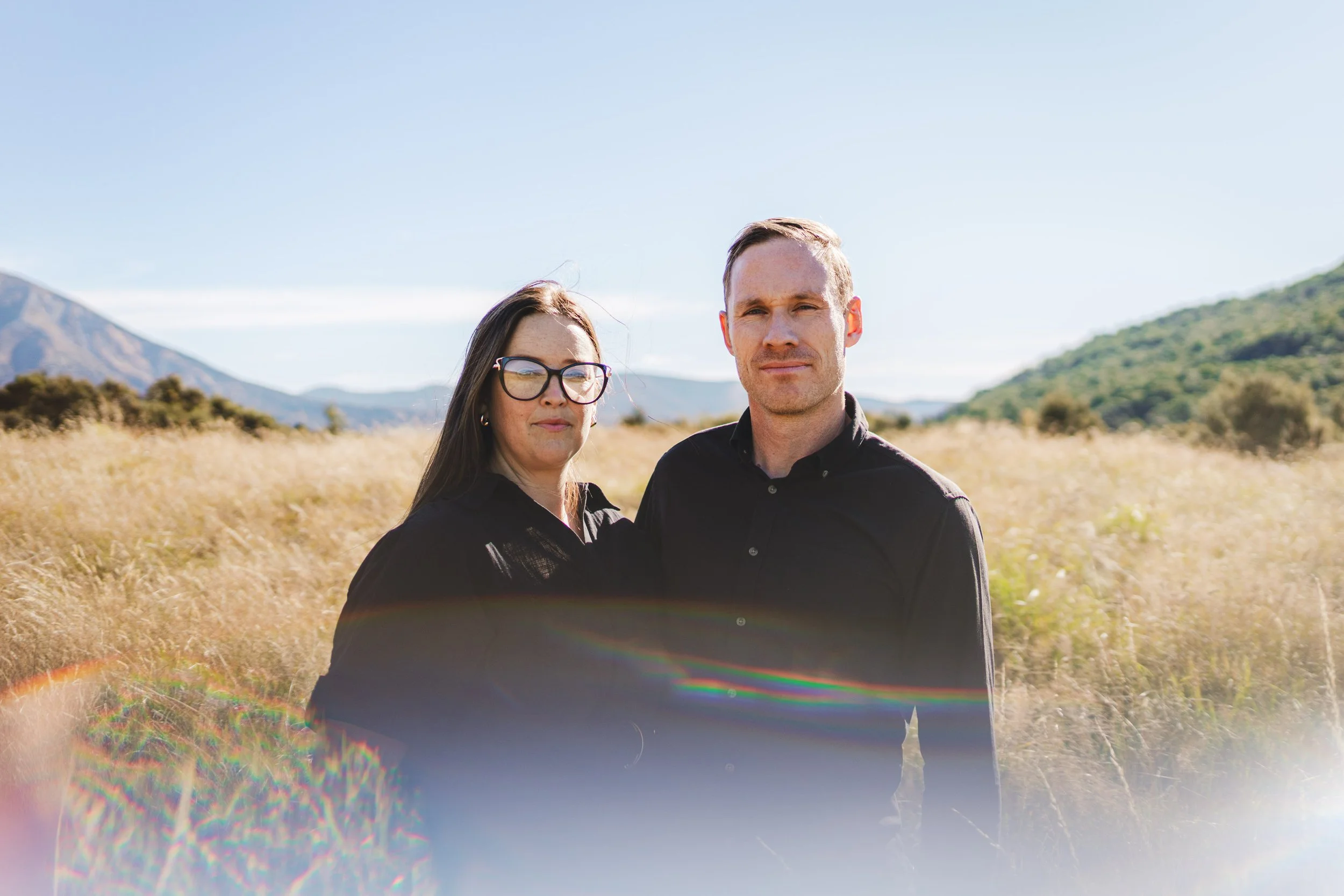 A man and a woman standing in a grassy field with mountains in the background, both wearing black shirts, during daytime.
