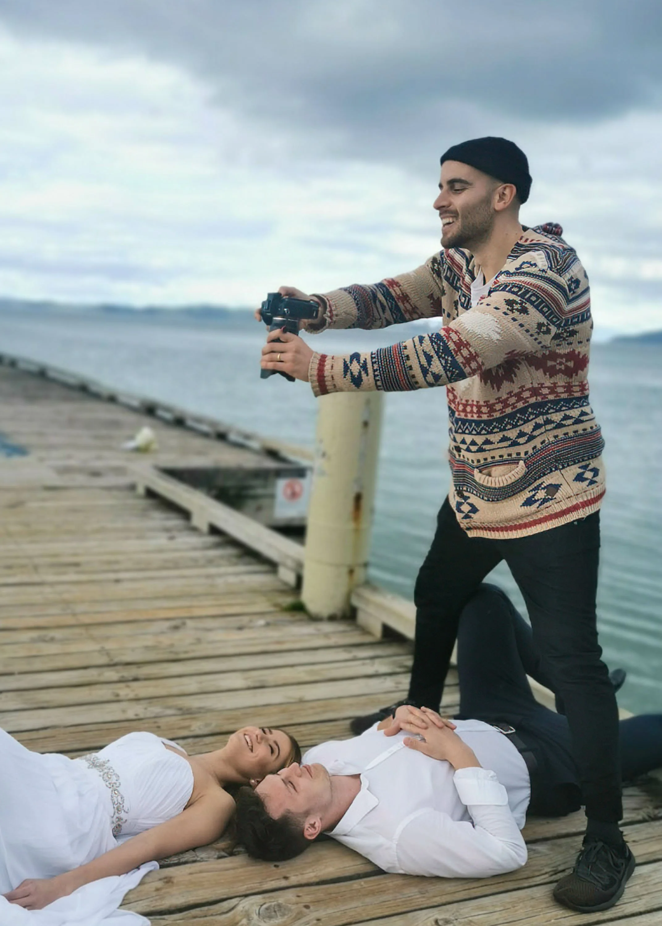 A man is taking a photo with a camera while two women lie on a wooden dock by the water. One woman is wearing a white dress and the other is in a white shirt. The scene appears joyful with both women smiling.