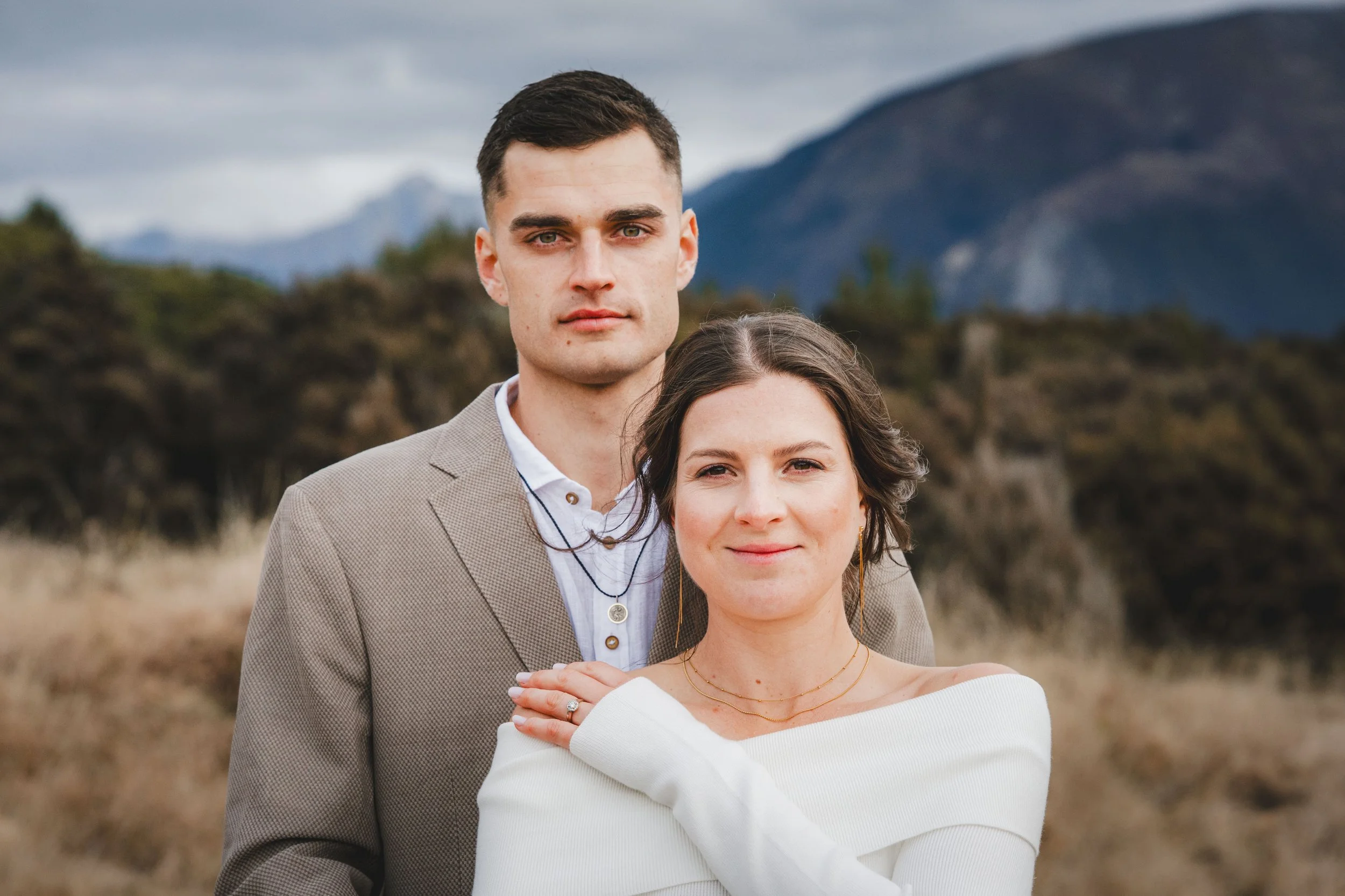 A man and woman standing outdoors with a mountainous landscape in the background. The man has dark hair, is wearing a beige blazer and a white shirt, and looks directly at the camera. The woman has curly brown hair, is wearing a white top, and is smi
